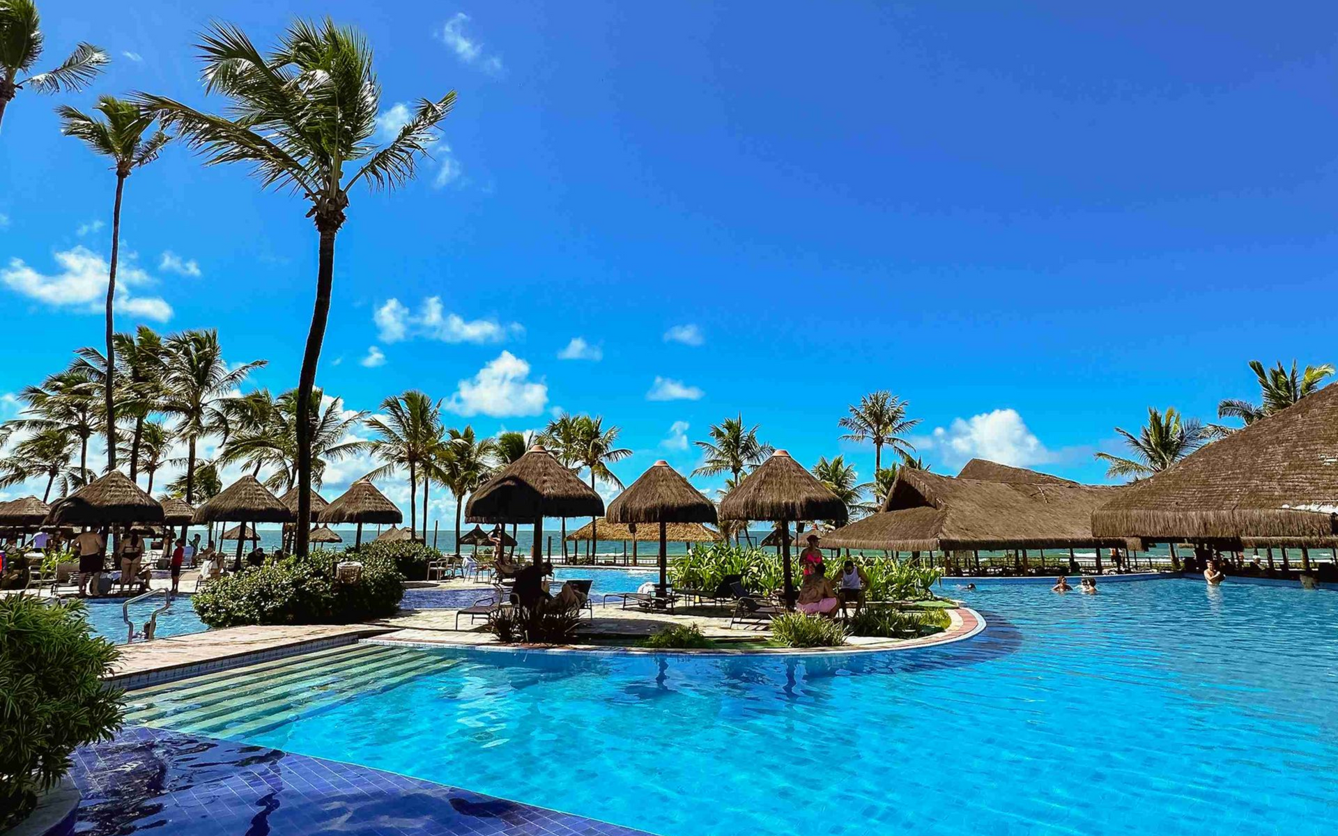 Piscina principal de azulejos azuis sob céu limpo, cercada por quiosques de palha e coqueiros no resort Enotel Porto de Galinhas