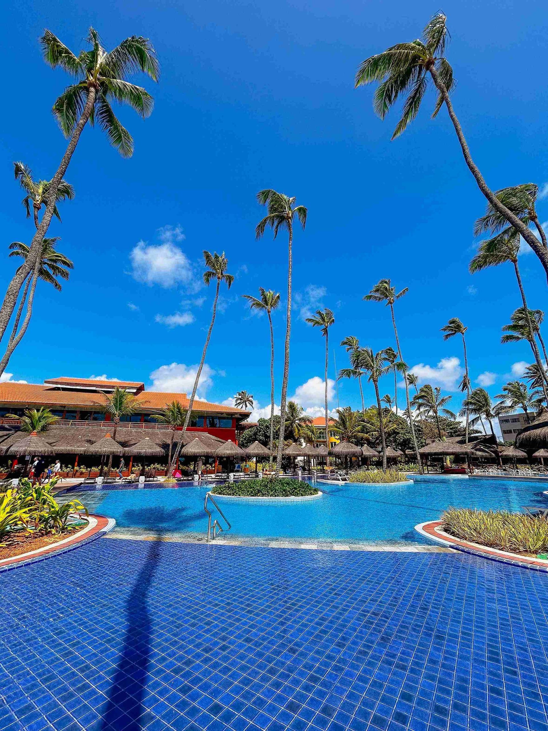 Amplo complexo de piscinas de azulejos azuis cercado por coqueiros altos e quiosques de palha no resort all inclusive Enotel Porto de Galinhas
