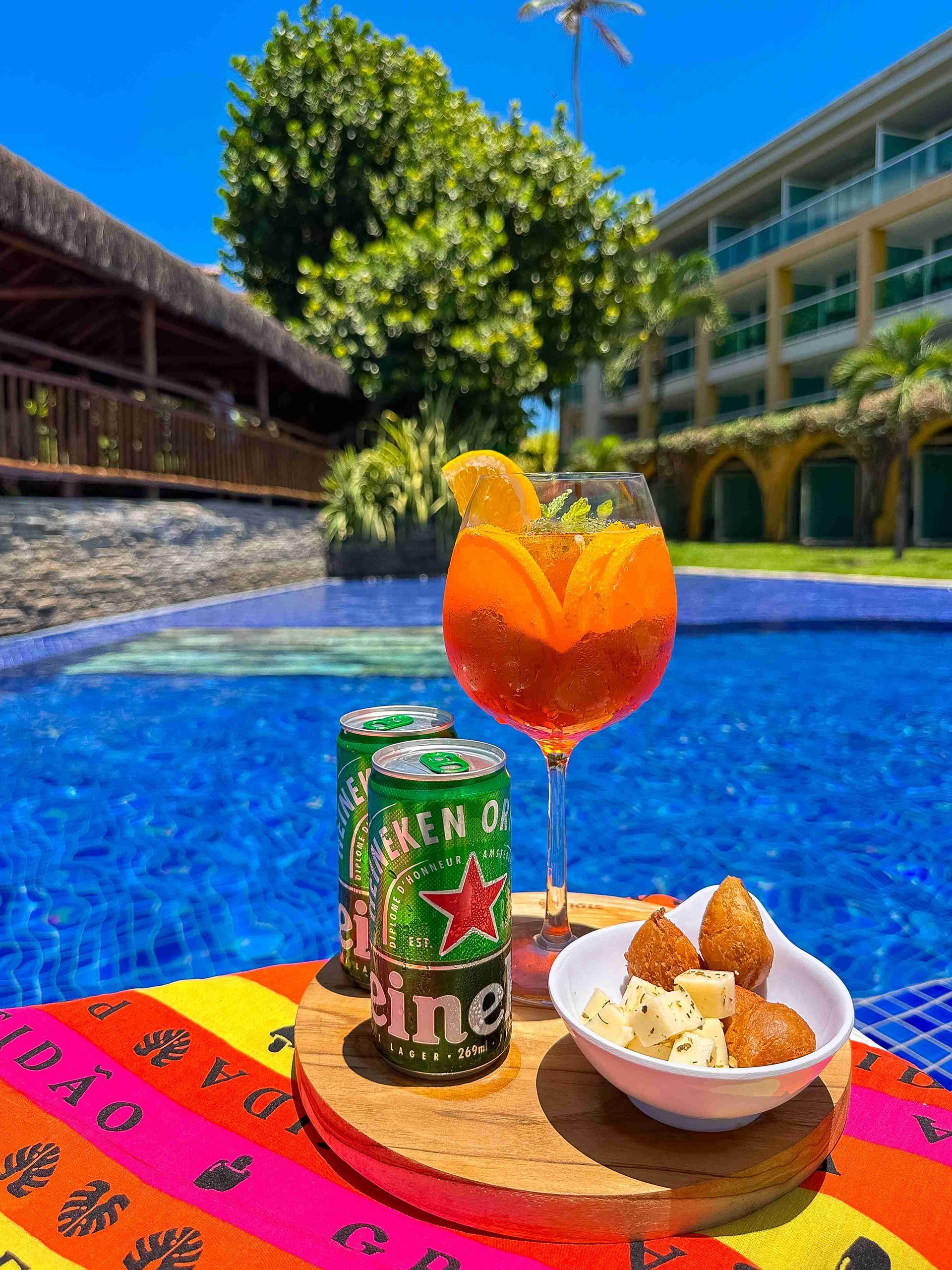 Taça de drink refrescante, latas de cerveja Heineken e petiscos de queijo servidos à beira da piscina azul no Enotel Porto de Galinhas