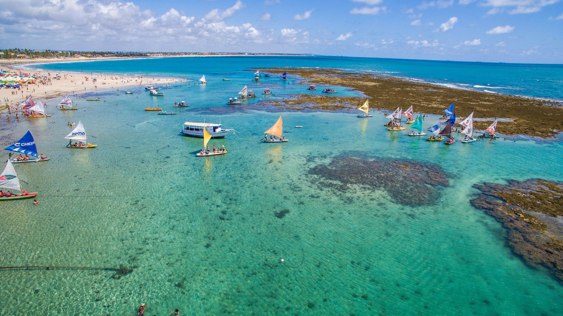 Vista aérea aproximada de jangadas coloridas navegando entre os recifes de corais e águas cristalinas de Porto de Galinhas, Pernambuco.