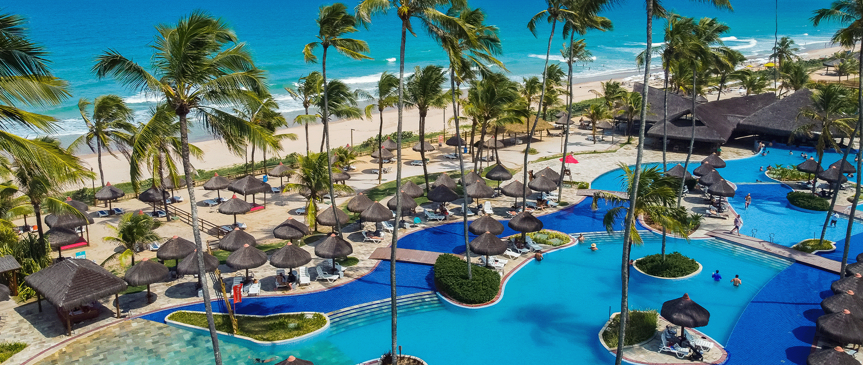 Vista aérea do Enotel Porto de Galinhas mostrando as extensas piscinas azuis e palhoças na praia de areia branca e mar turquesa, em dia ensolarado no resort all inclusive.
