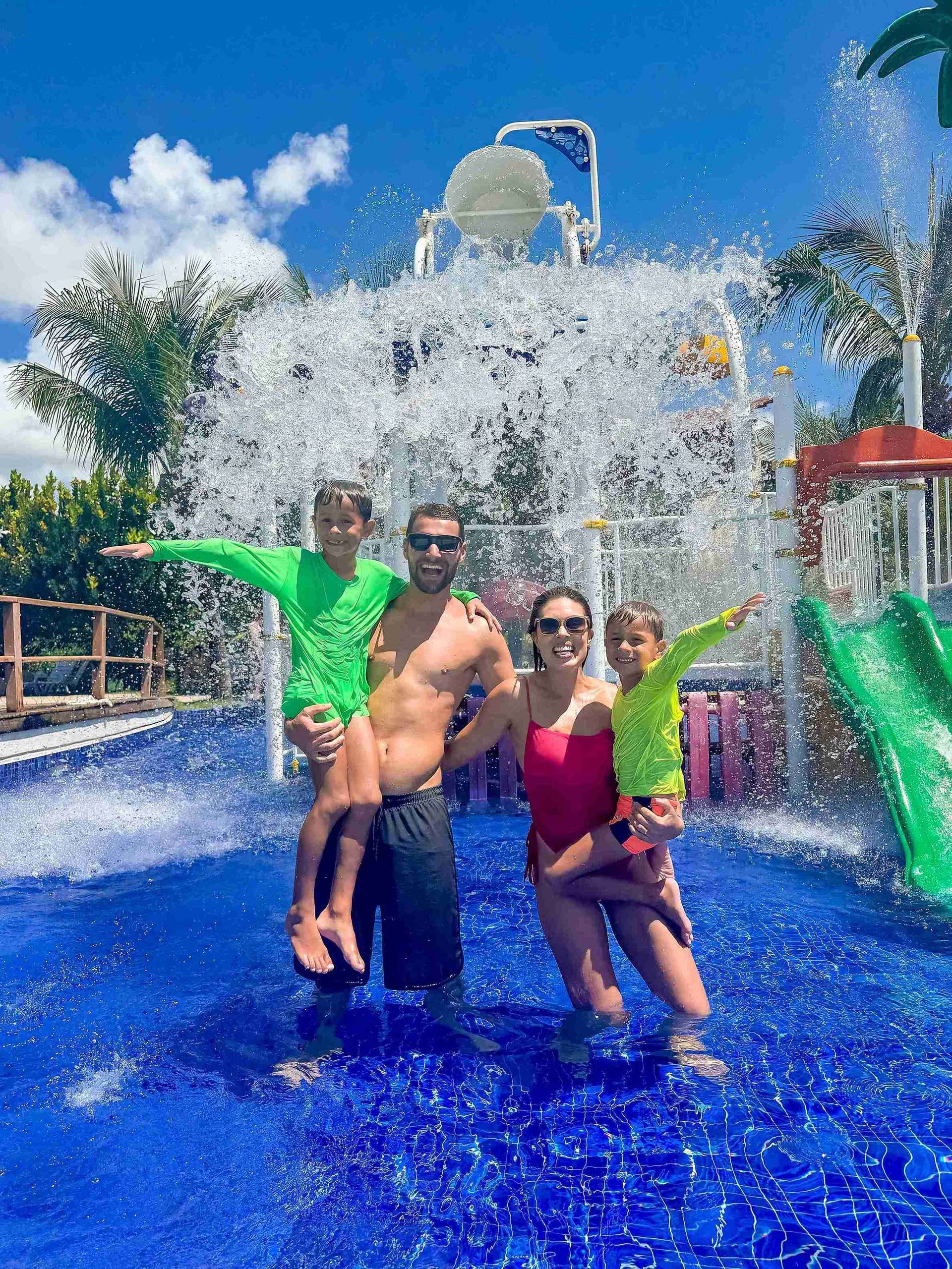 Família sorridente se divertindo sob grande cascata de água no parque aquático infantil do resort all inclusive Enotel Porto de Galinhas