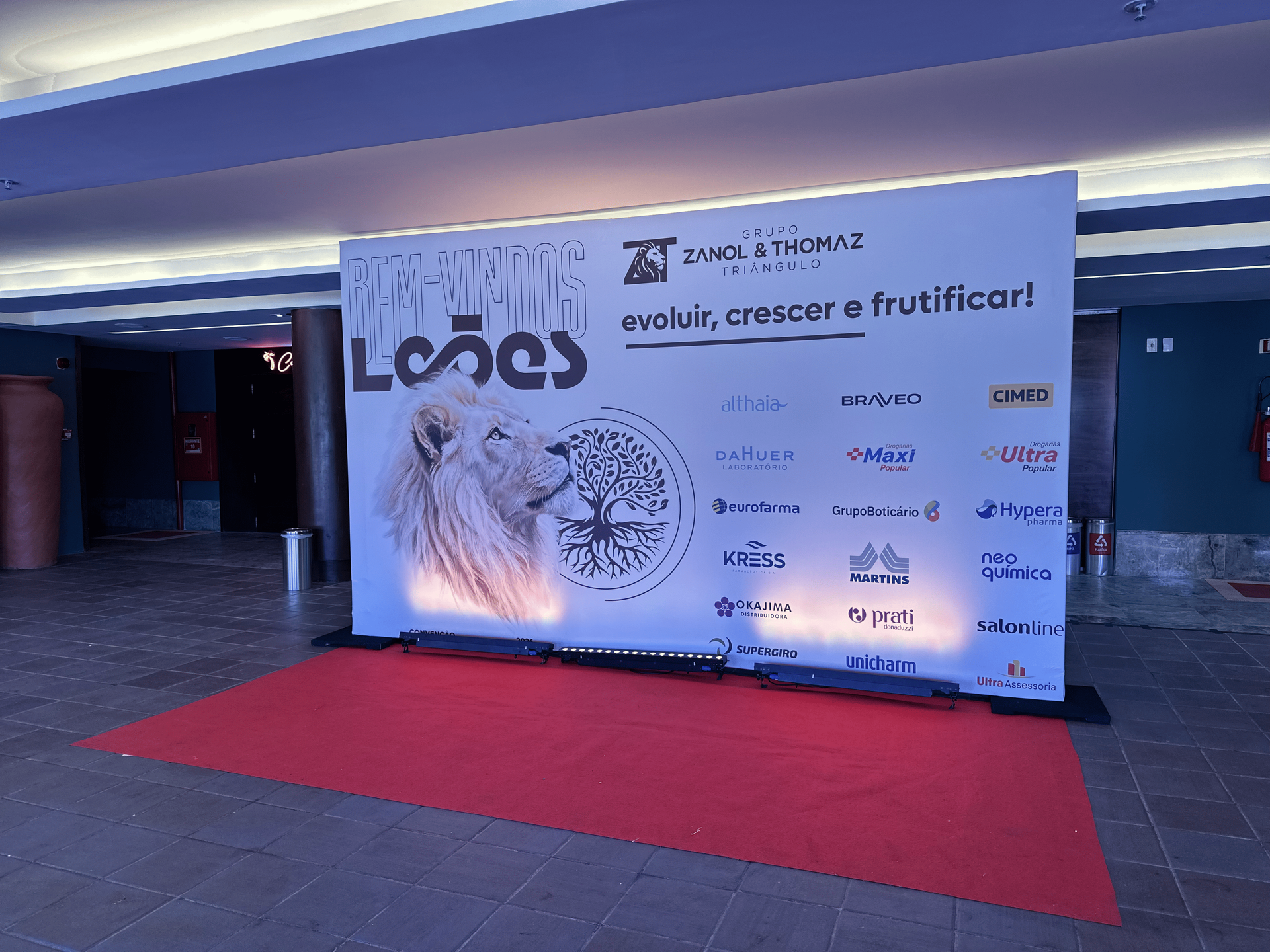 Painel backdrop de boas-vindas para convenção com estampa de leão e logotipos de patrocinadores posicionado sobre tapete vermelho no Foyer Brennand