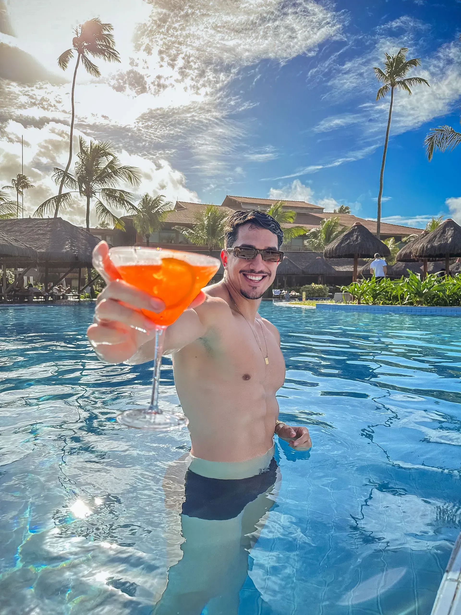 Homem sorridente segurando um drink refrescante dentro da piscina principal do Enotel Porto de Galinhas em um dia ensolarado