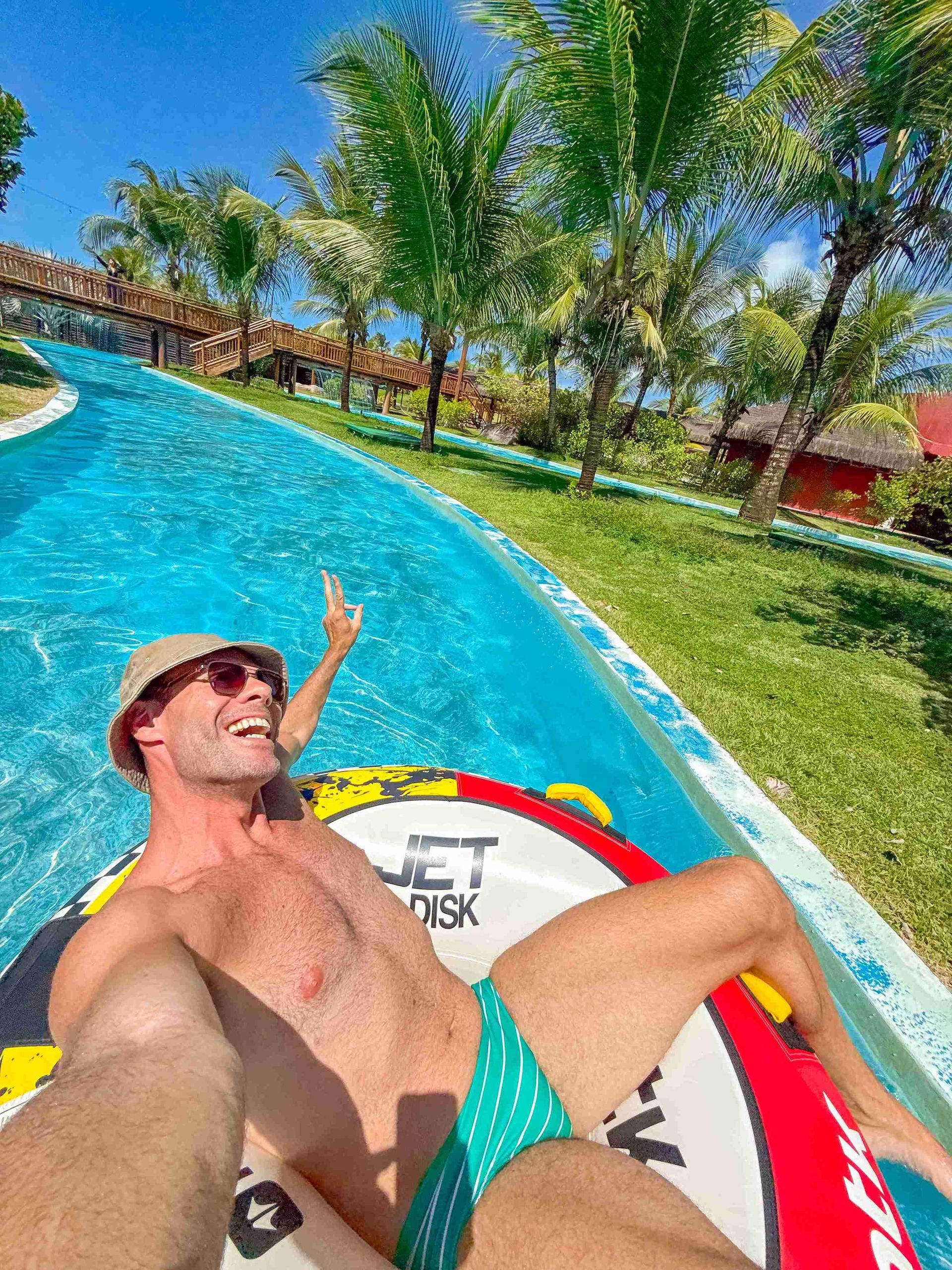 Homem sorridente de chapéu relaxando em uma boia inflável no rio lento do Enotel Porto de Galinhas, cercado por coqueiros e áreas verdes do resort