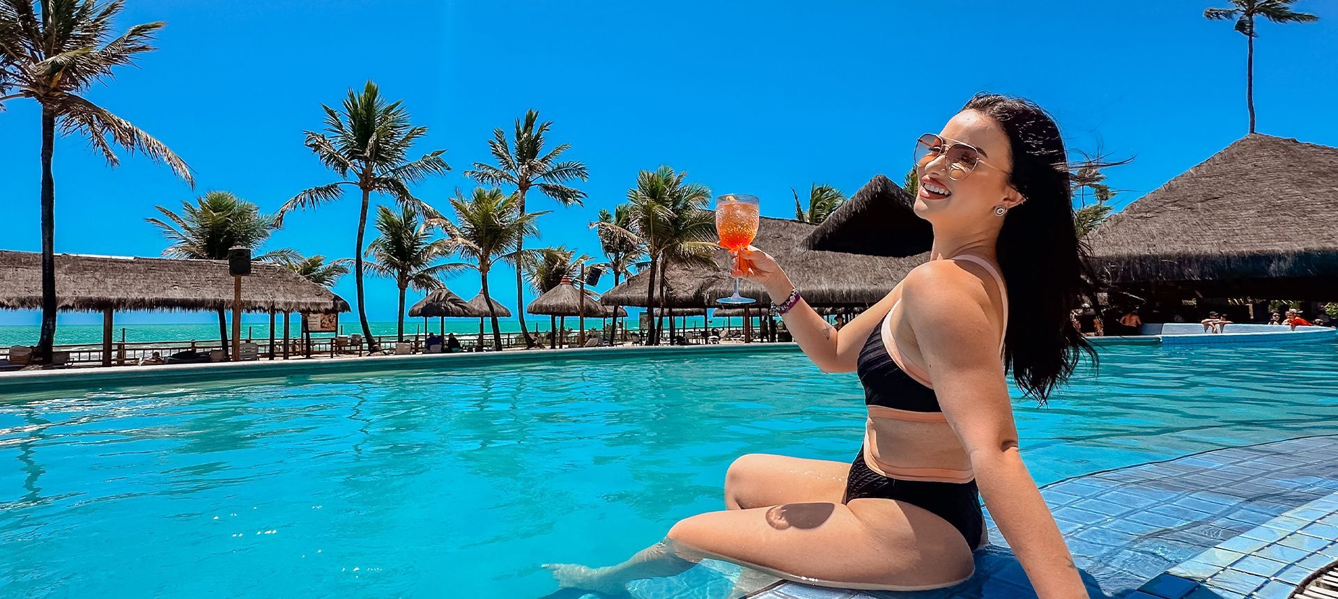 Mulher sorridente de óculos escuros relaxando na piscina principal com um drink refrescante na mão no Enotel Porto de Galinhas