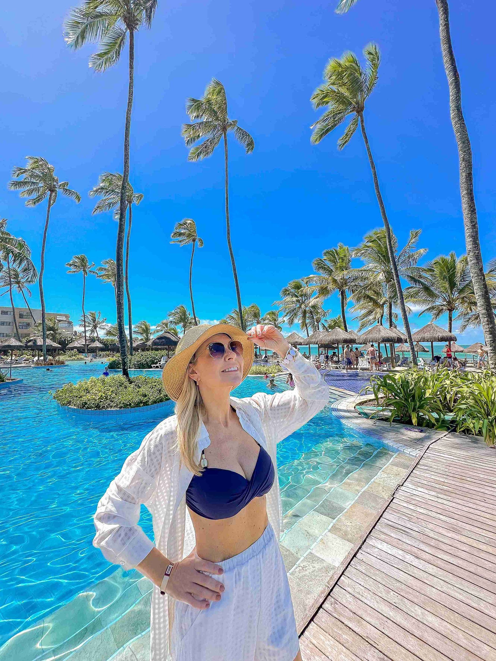 Mulher de chapéu de palha sorrindo à beira da grande piscina azul cercada por coqueiros altos no resort de praia Enotel Porto de Galinhas