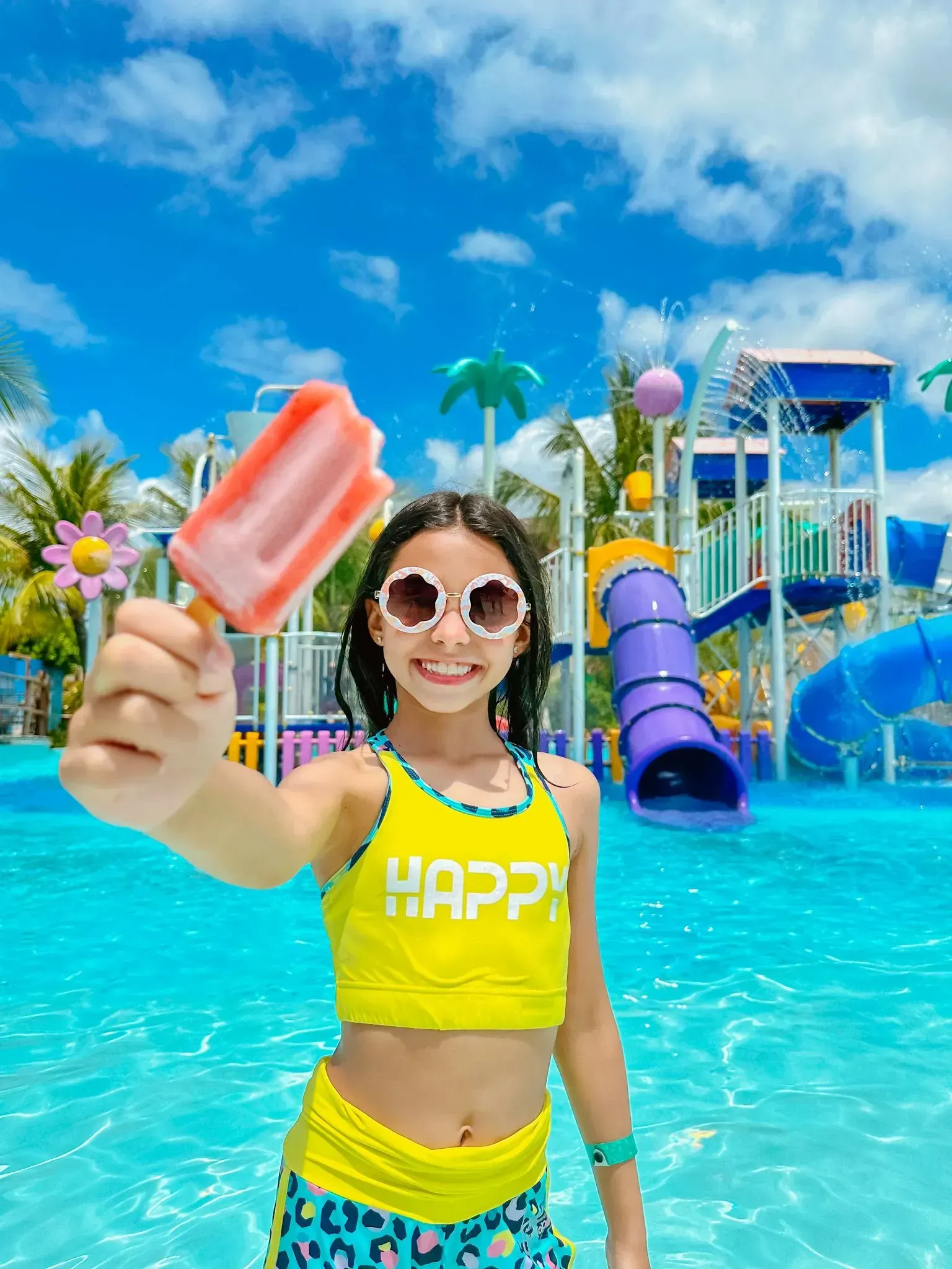 Garota sorridente de óculos escuros segurando um picolé em frente às atrações e toboáguas do parque aquático do Enotel Porto de Galinhas