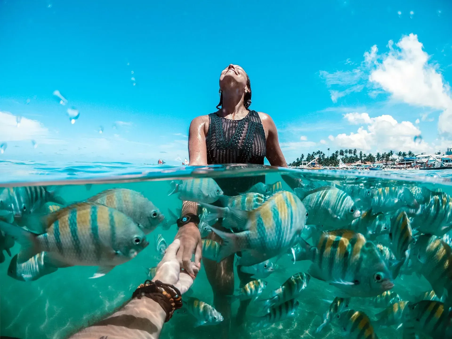 Mulher sorridente de mãos dadas cercada por peixes sargentinhos em águas cristalinas durante passeio nas piscinas naturais de Porto de Galinhas