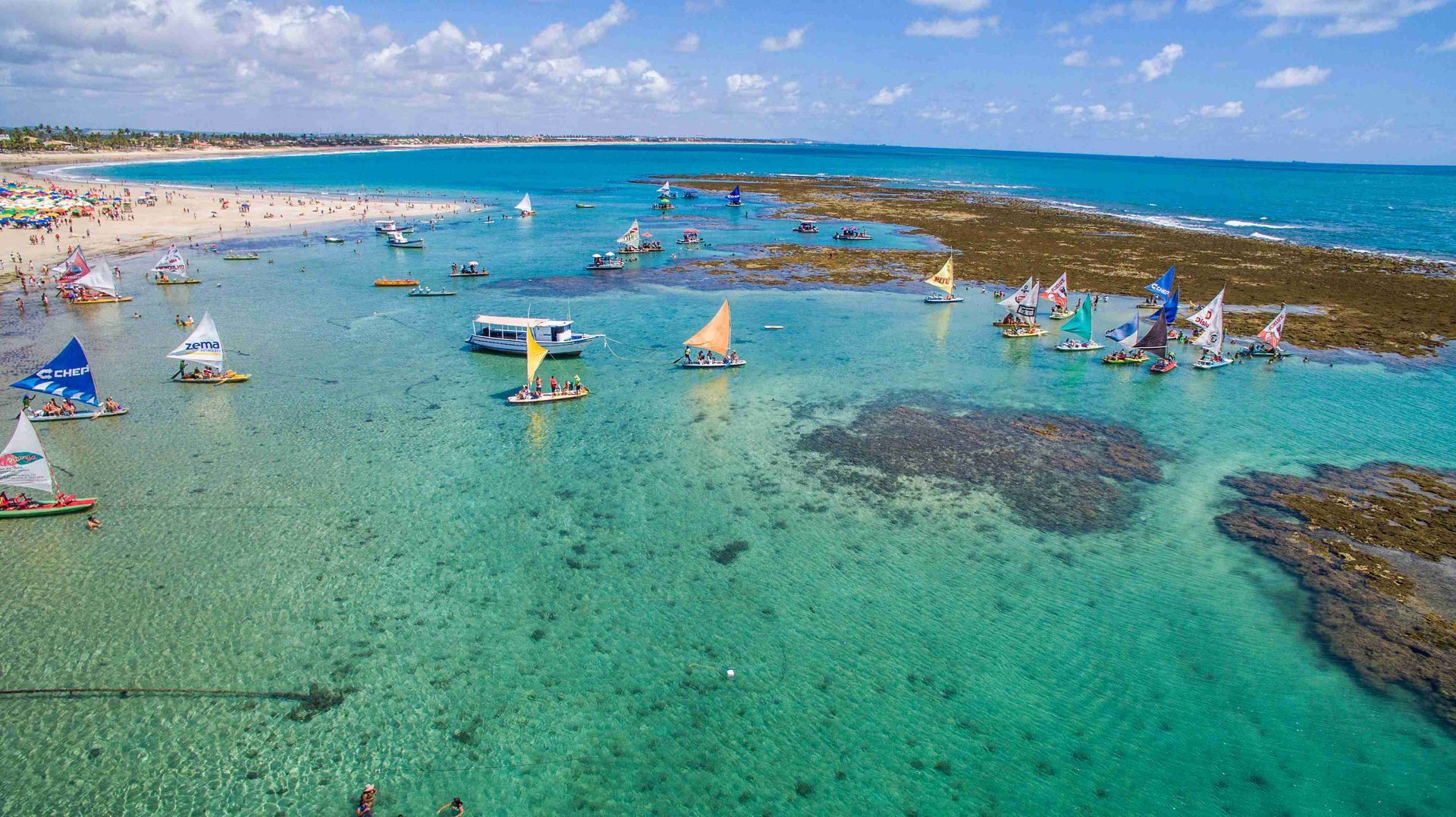 Vista aérea das piscinas naturais de Porto de Galinhas com águas cristalinas e jangadas coloridas navegando perto dos recifes de corais