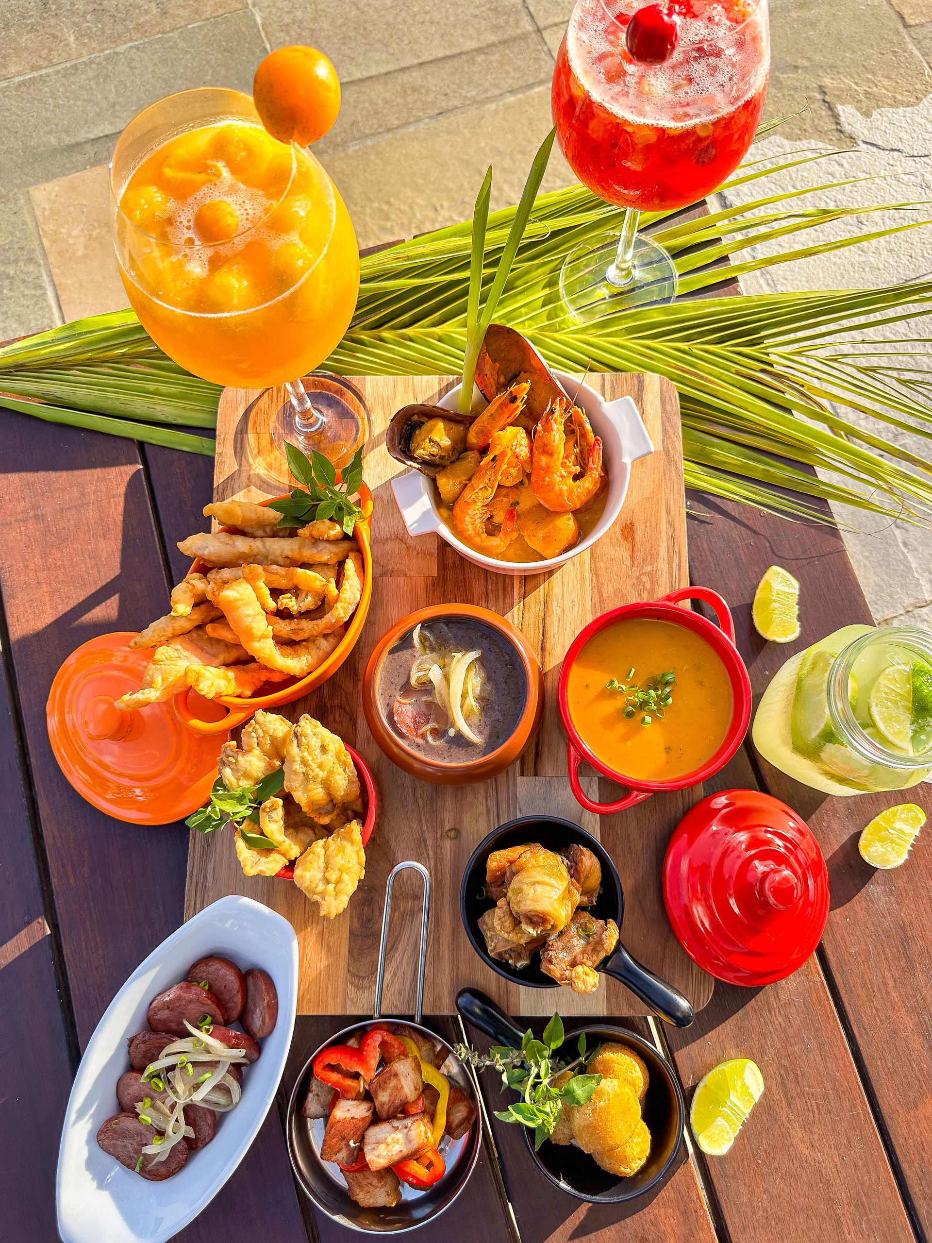 Vista aérea de uma mesa de madeira com uma tábua repleta de petiscos brasileiros: caldinho de feijão, moqueca de camarão com mexilhões, peixe frito, linguiça acebolada, torresmo e bolinhos. A composição inclui dois drinks vibrantes em taças grandes (um laranja e um vermelho), uma jarra de caipirinha com limão e decoração com folha de palmeira.