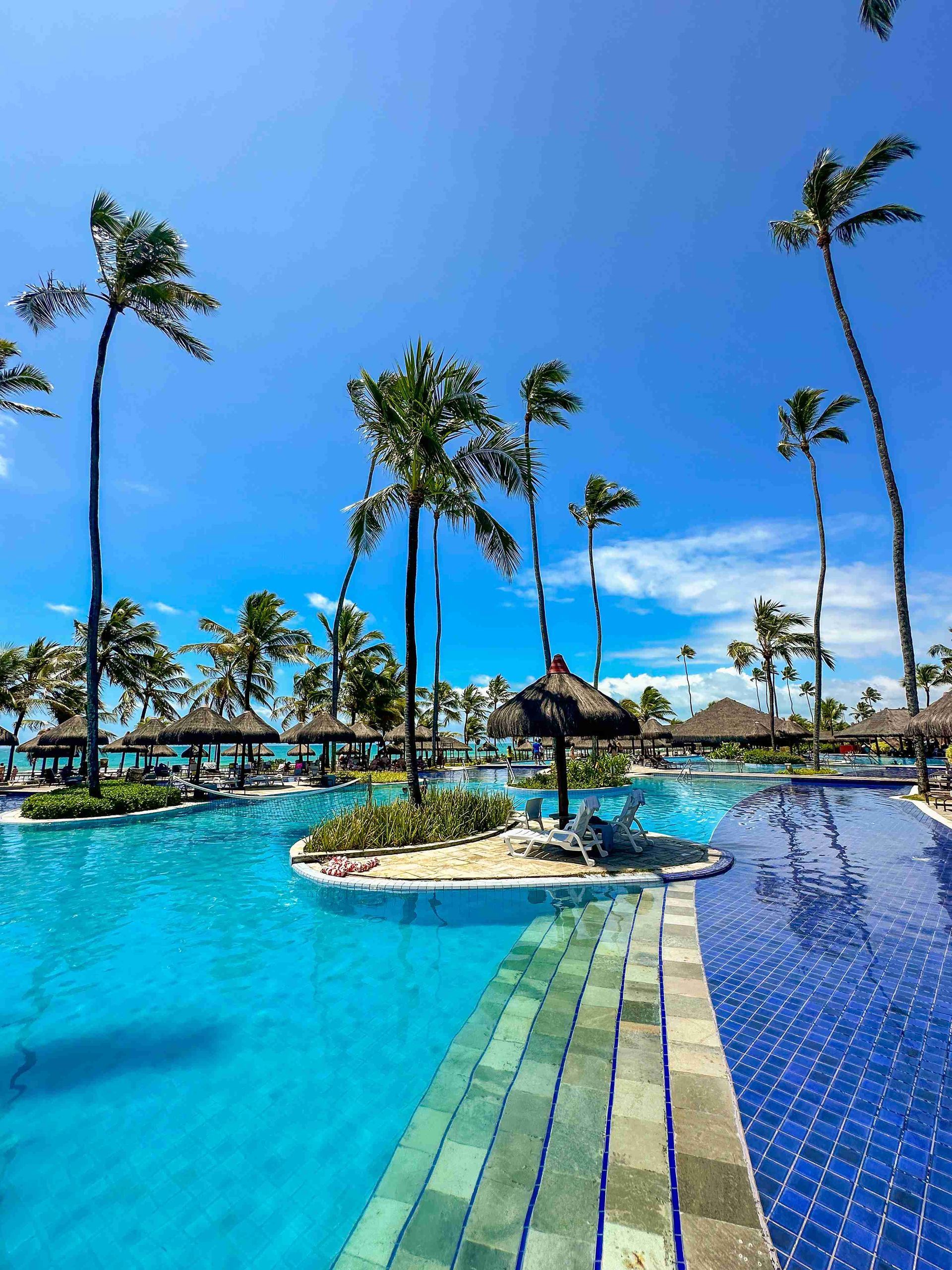 Vista vertical da piscina principal de águas claras com ilhas de coqueiros, quiosques de palha e espreguiçadeiras no resort all inclusive Enotel Porto de Galinhas