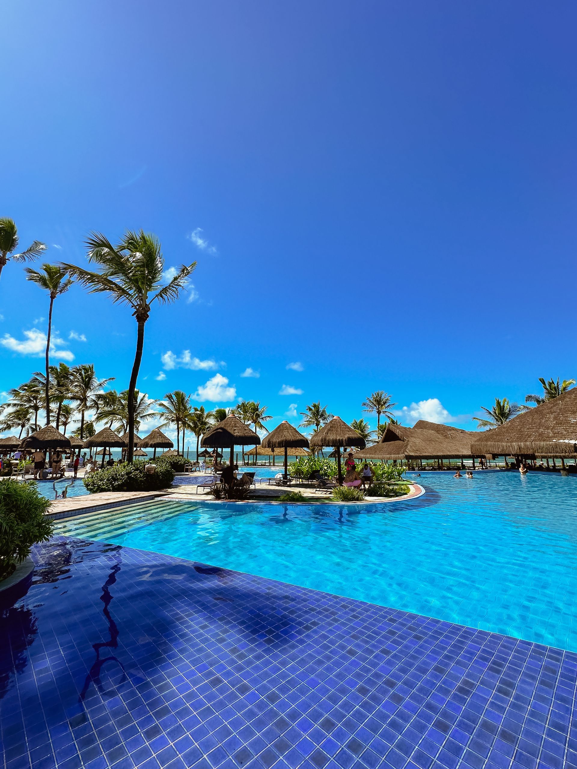 Ampla piscina de azulejos azuis cercada por quiosques de palha, coqueiros altos e áreas de relaxamento sob céu ensolarado no resort Enotel Porto de Galinhas