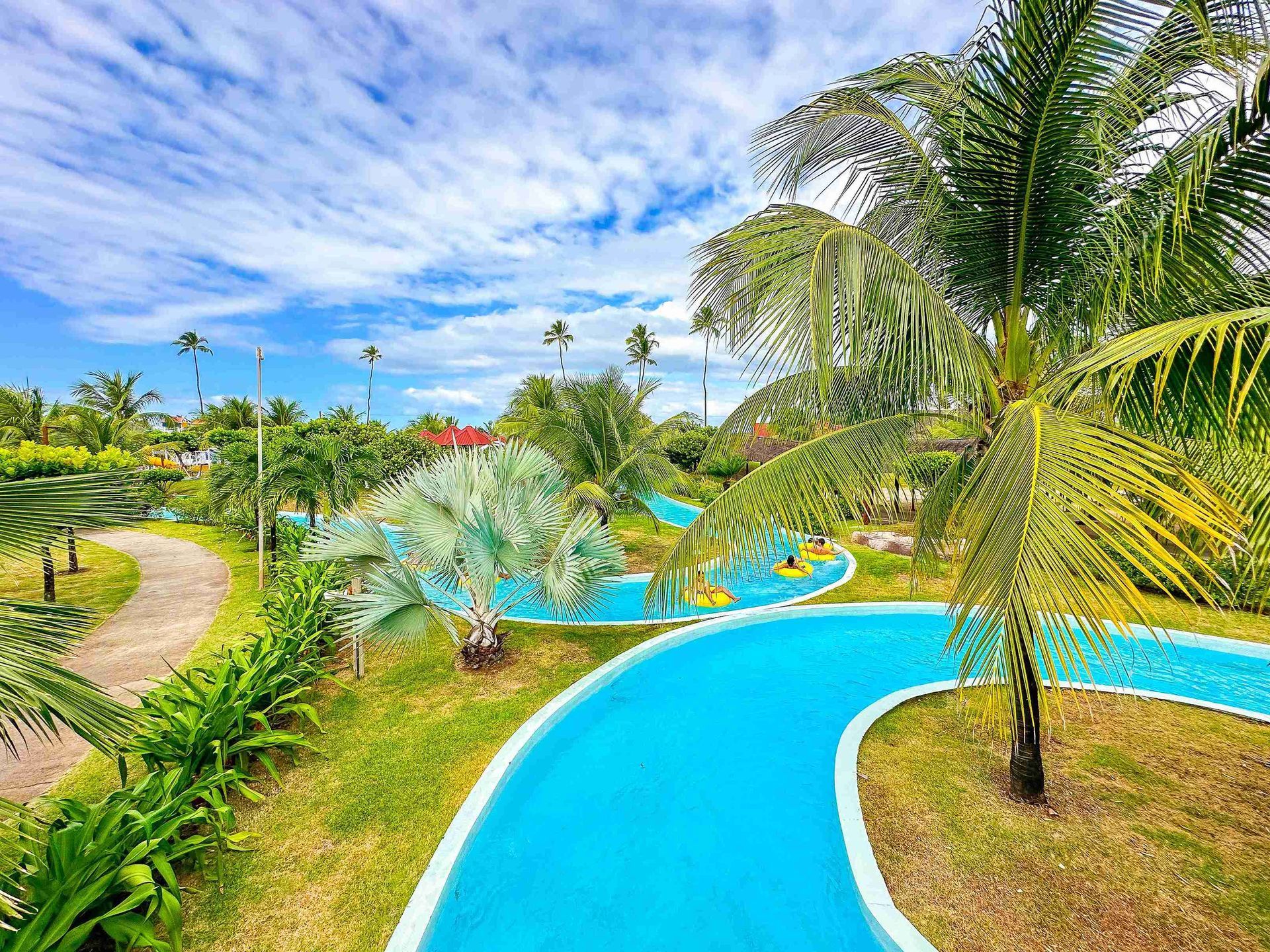 Vista do circuito do rio lento com águas turquesas, cercado por coqueiros, palmeira-azul e gramado, exibindo hóspedes em boias amarelas no parque aquático do Enotel Porto de Galinhas