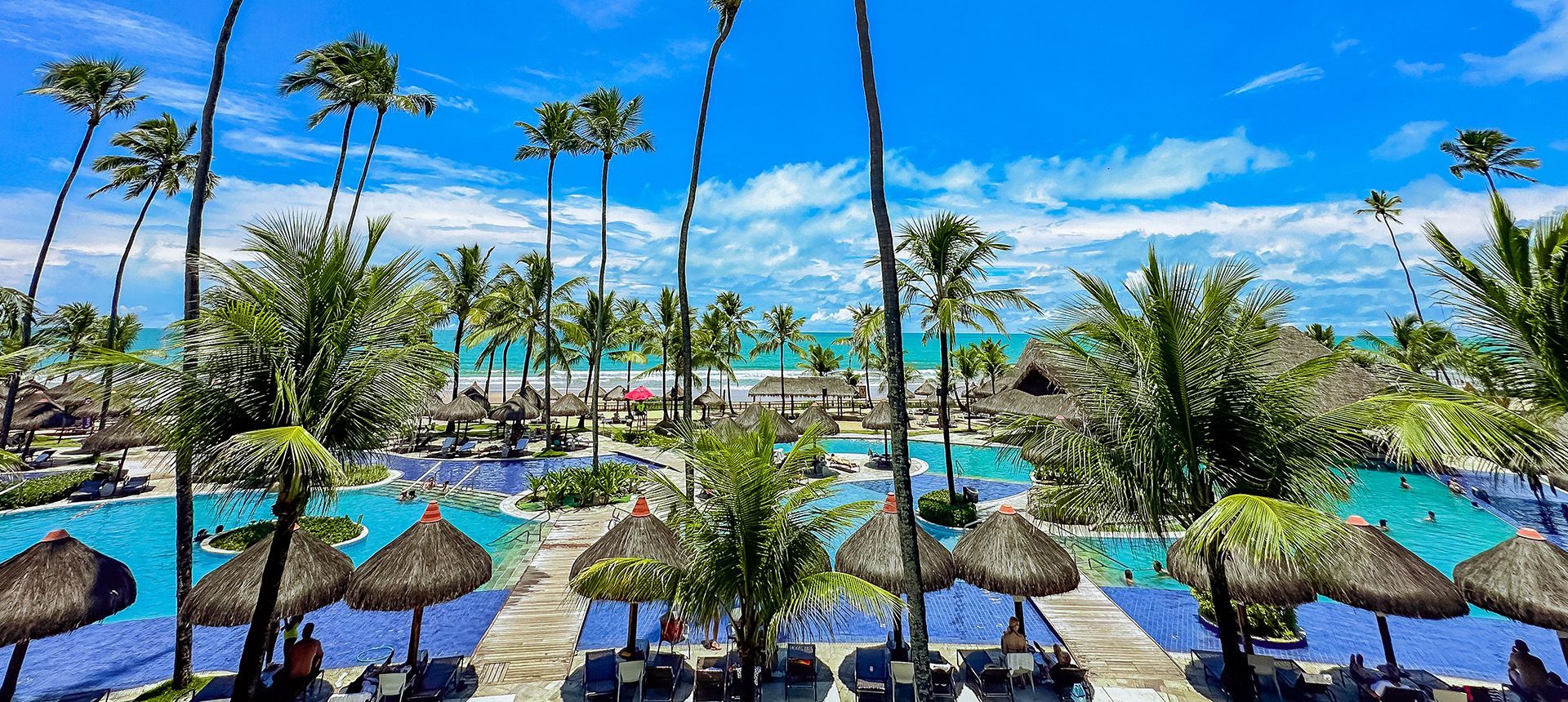Vista panorâmica elevada mostrando a grandiosa estrutura de piscinas tropicais e quiosques de palha de frente para a praia no Enotel Porto de Galinhas