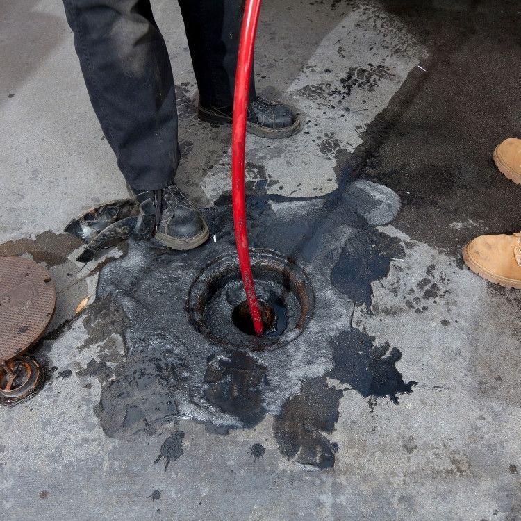 Red hose draining a manhole in wet pavement beside workers’ boots and spilled dark liquid.