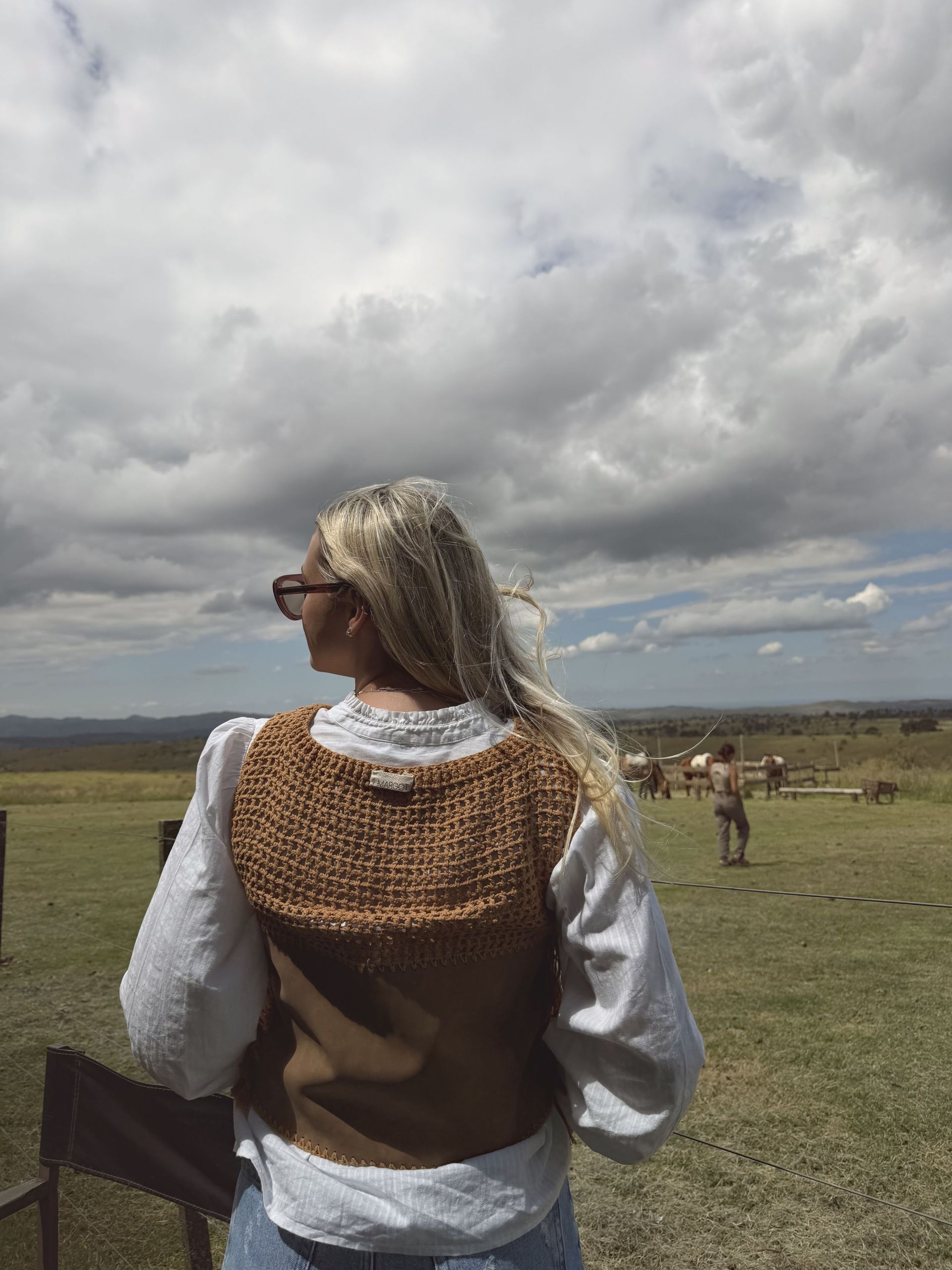 Mujer con gafas de sol y chaleco marrón, mirando hacia otro lado, en una habitación rústica.