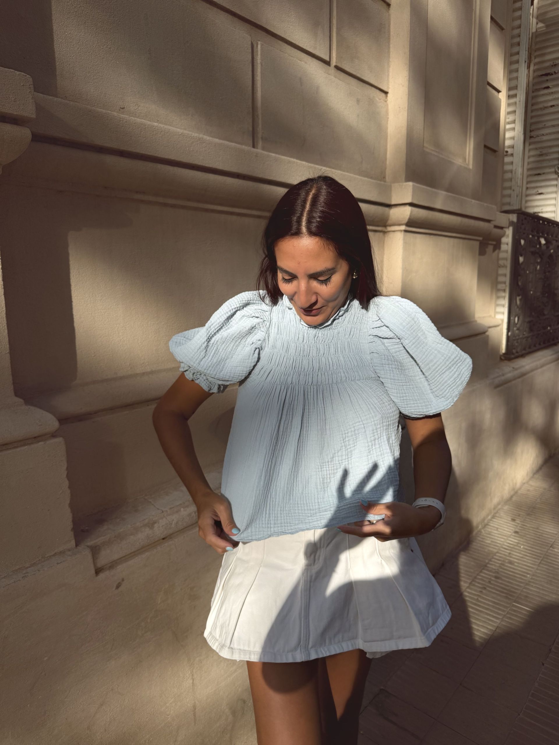 Una mujer con una blusa azul claro con mangas abullonadas y una falda blanca sonríe cerca de un edificio de color canela.