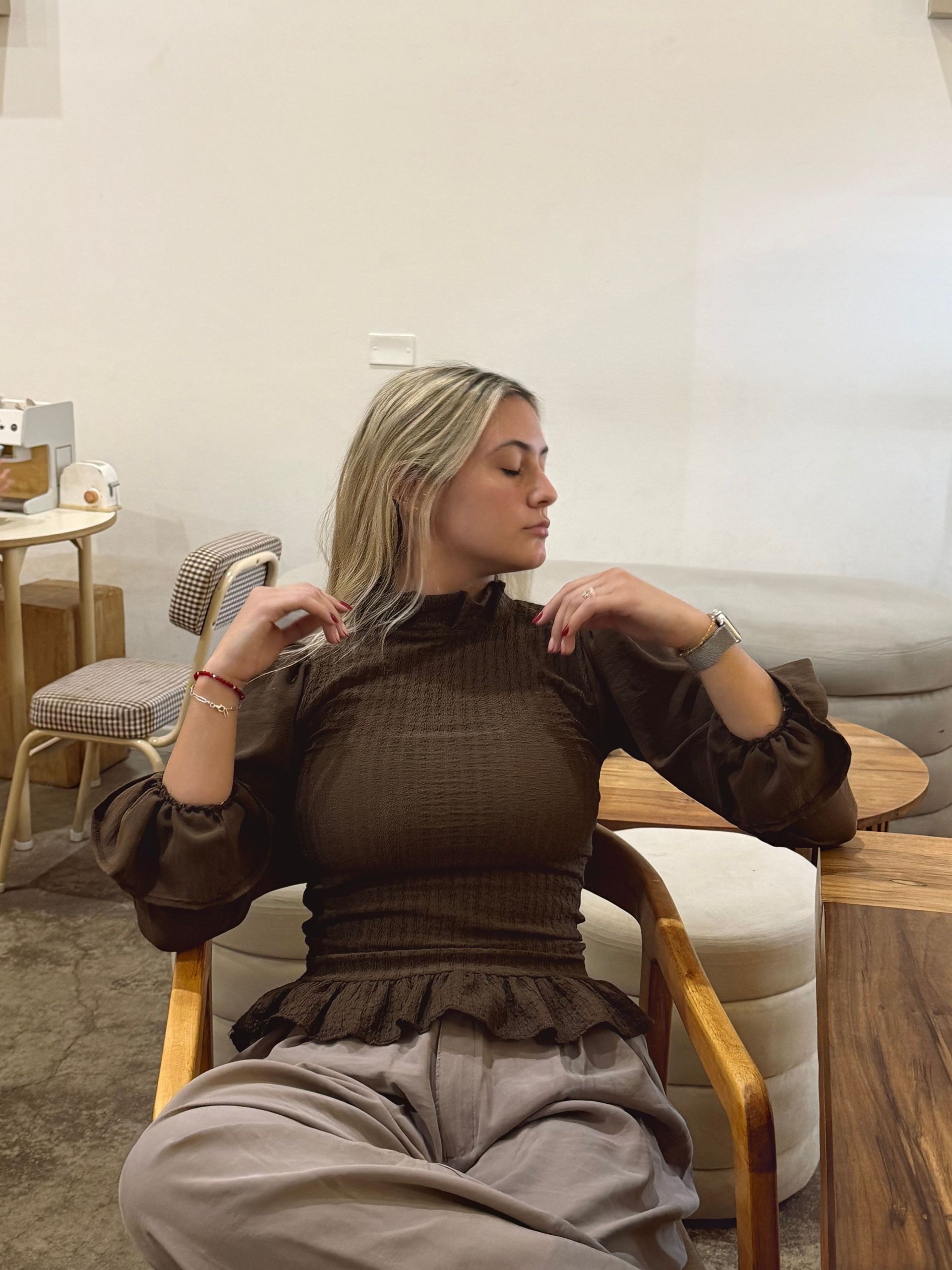 Mujer con blusa marrón sentada, estirando el cuello. Manos en alto y cabeza inclinada, en un espacio interior.