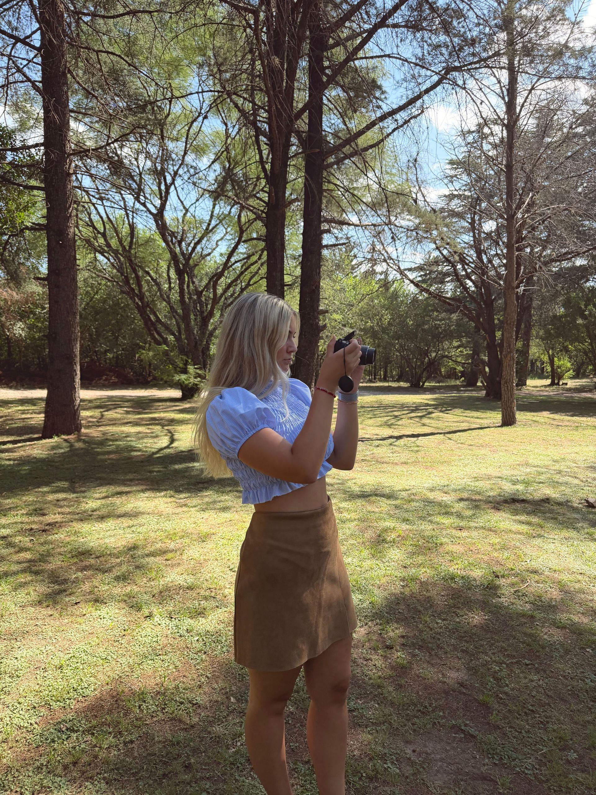 Mujer con top azul y falda marrón sonríe al aire libre, rodeada de árboles.