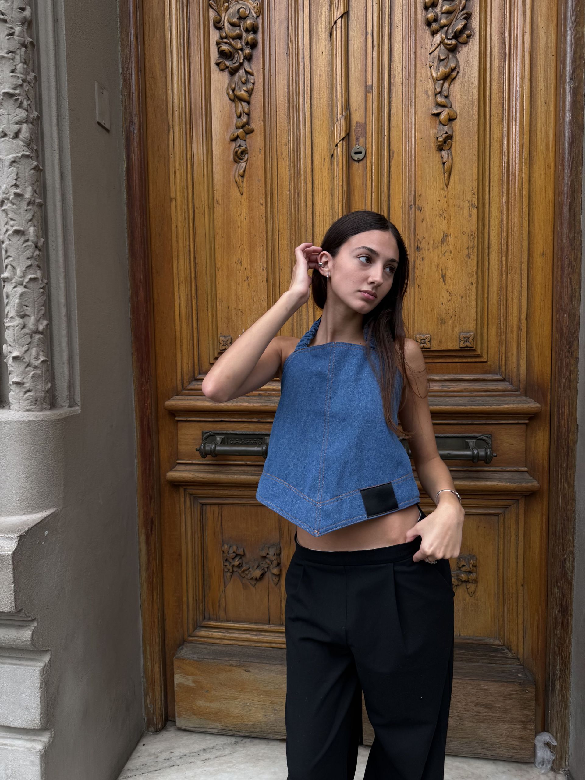 Mujer con top vaquero y pantalón negro posa frente a una puerta de madera.