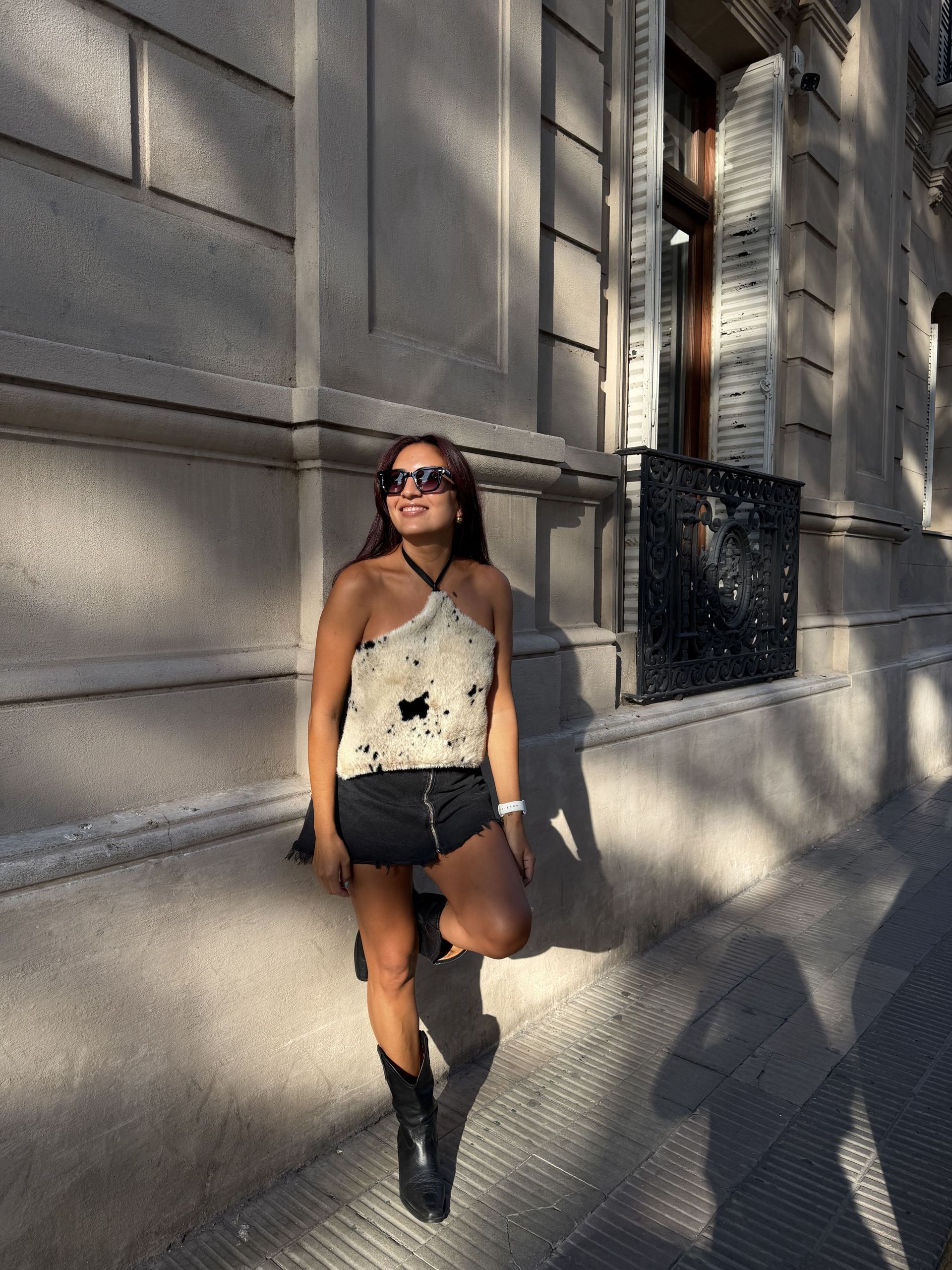 Una mujer con gafas de sol y un top con cuello halter se apoya en un edificio bajo la luz del sol.