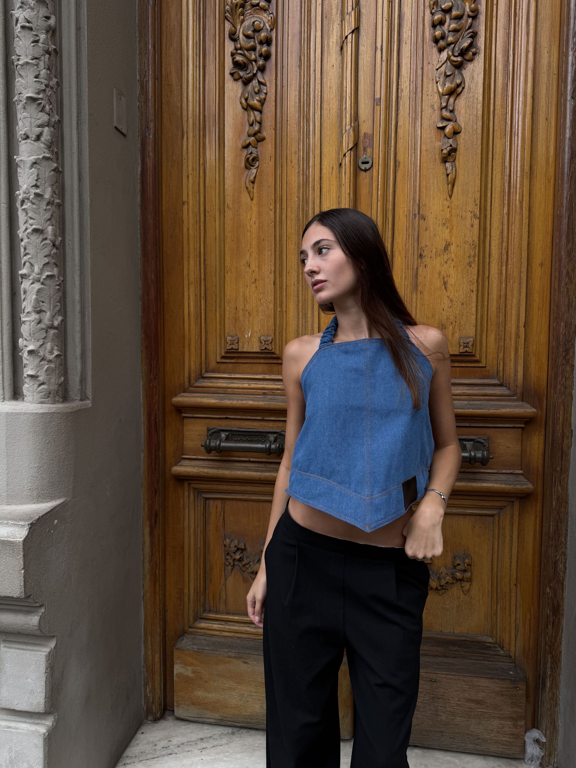 Mujer con top vaquero y pantalón negro posa frente a una puerta de madera.