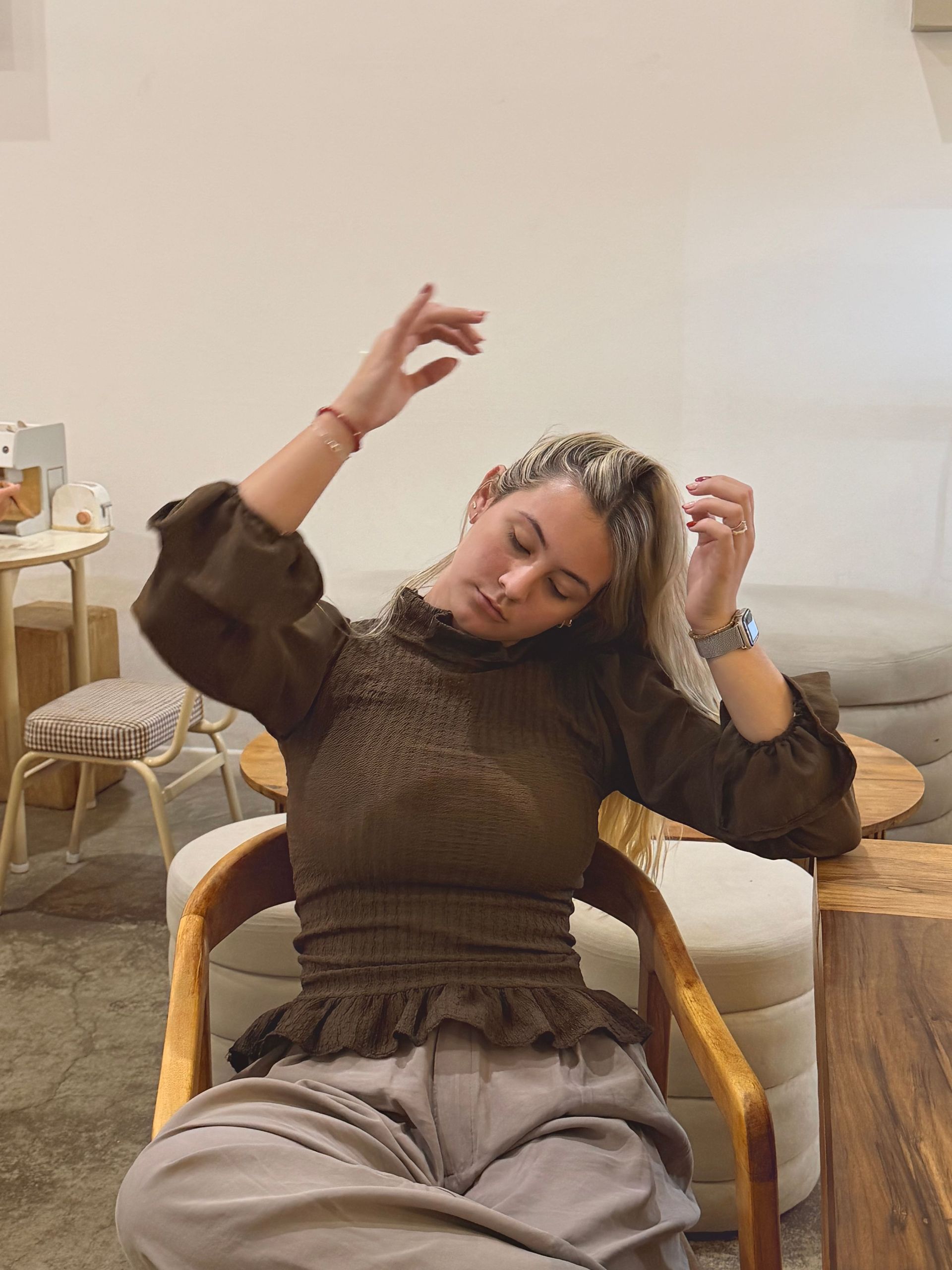 Mujer con blusa marrón sentada, estirando el cuello. Manos en alto y cabeza inclinada, en un espacio interior.