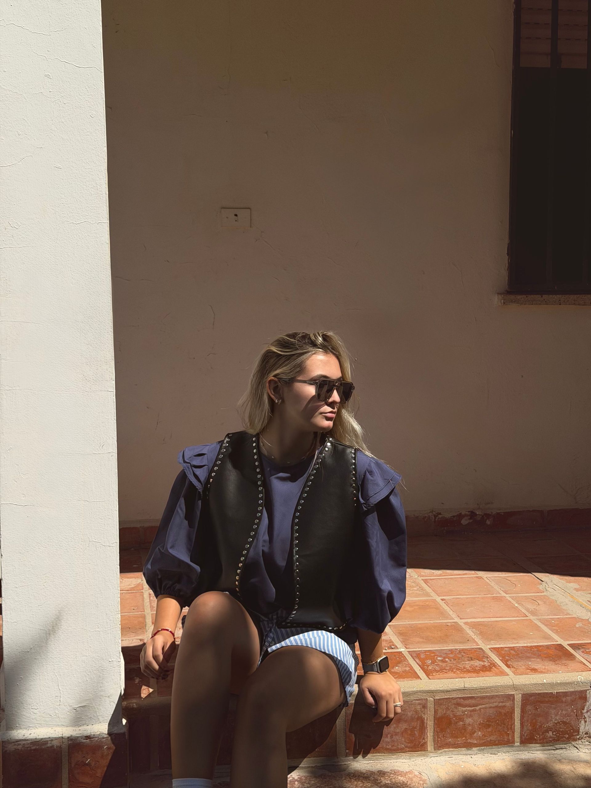 Mujer con gafas de sol, chaleco y pantalones cortos, sentada en unos escalones de ladrillo.