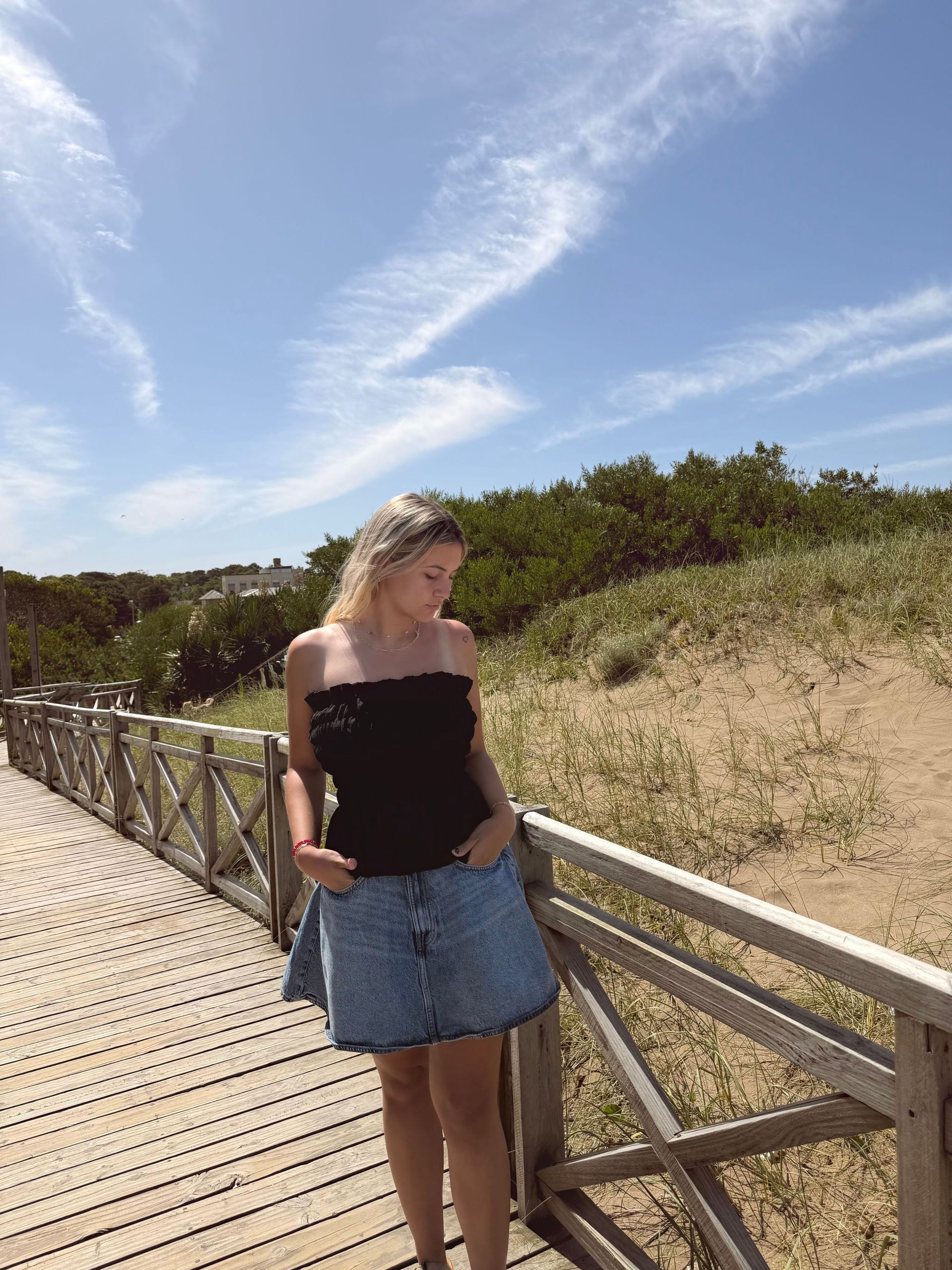 Persona rubia con top negro y shorts vaqueros, de pie sobre una pasarela de madera