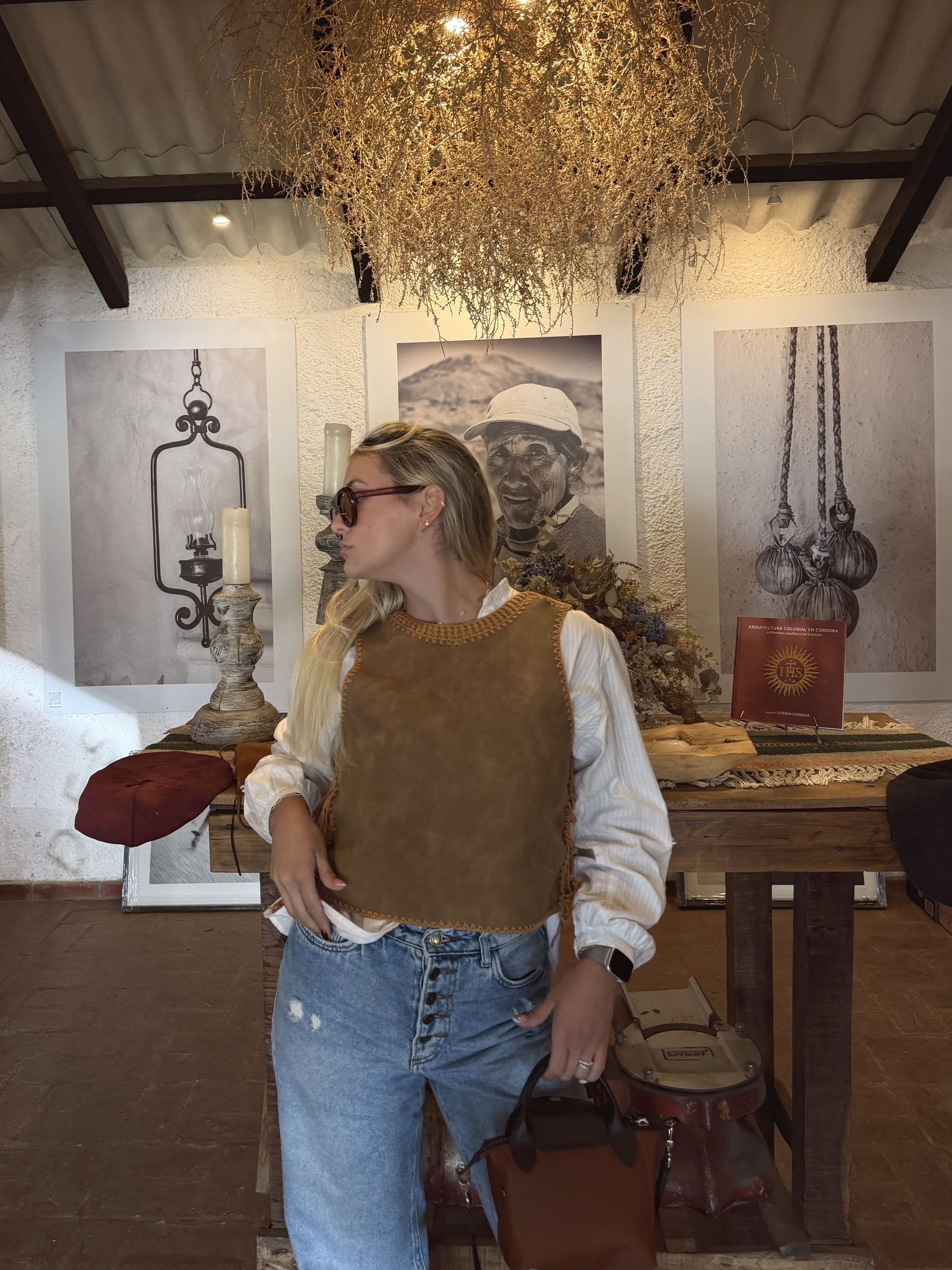 Mujer con gafas de sol y chaleco marrón, mirando hacia otro lado, en una habitación rústica.