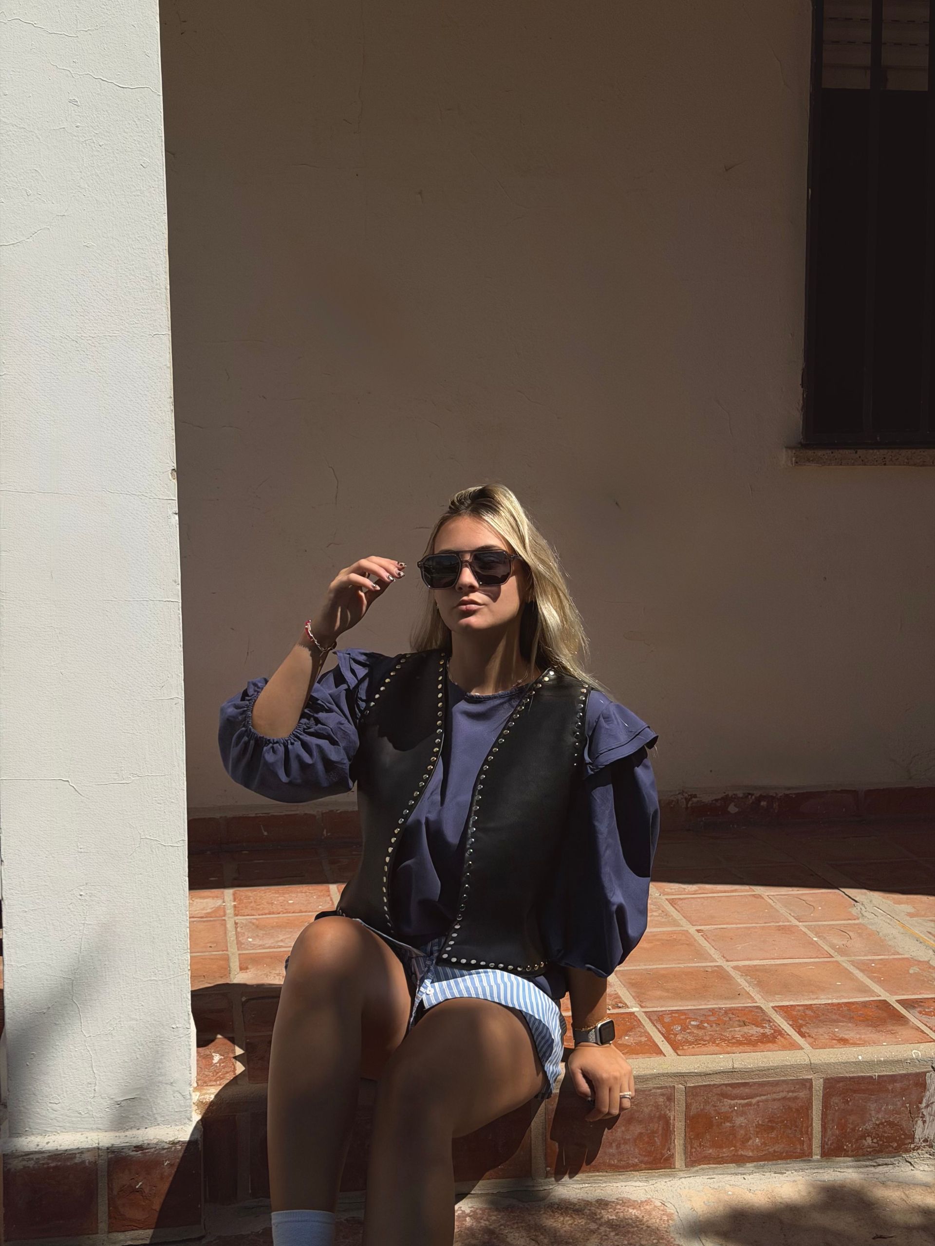 Mujer con gafas de sol, chaleco y pantalones cortos, sentada en unos escalones de ladrillo.