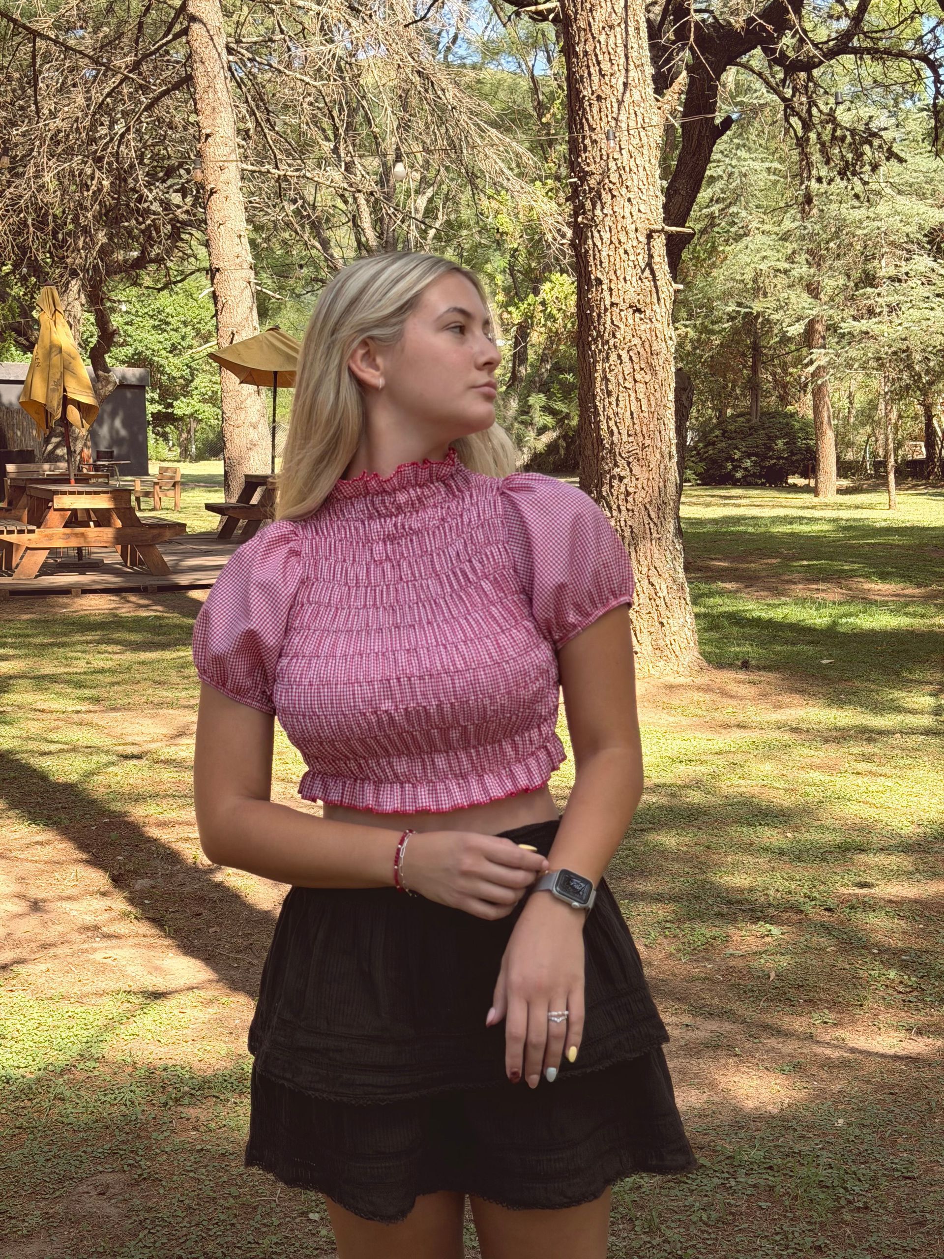 Mujer con top rosa y falda negra al aire libre, mirando a la izquierda. Árboles y vegetación al fondo.