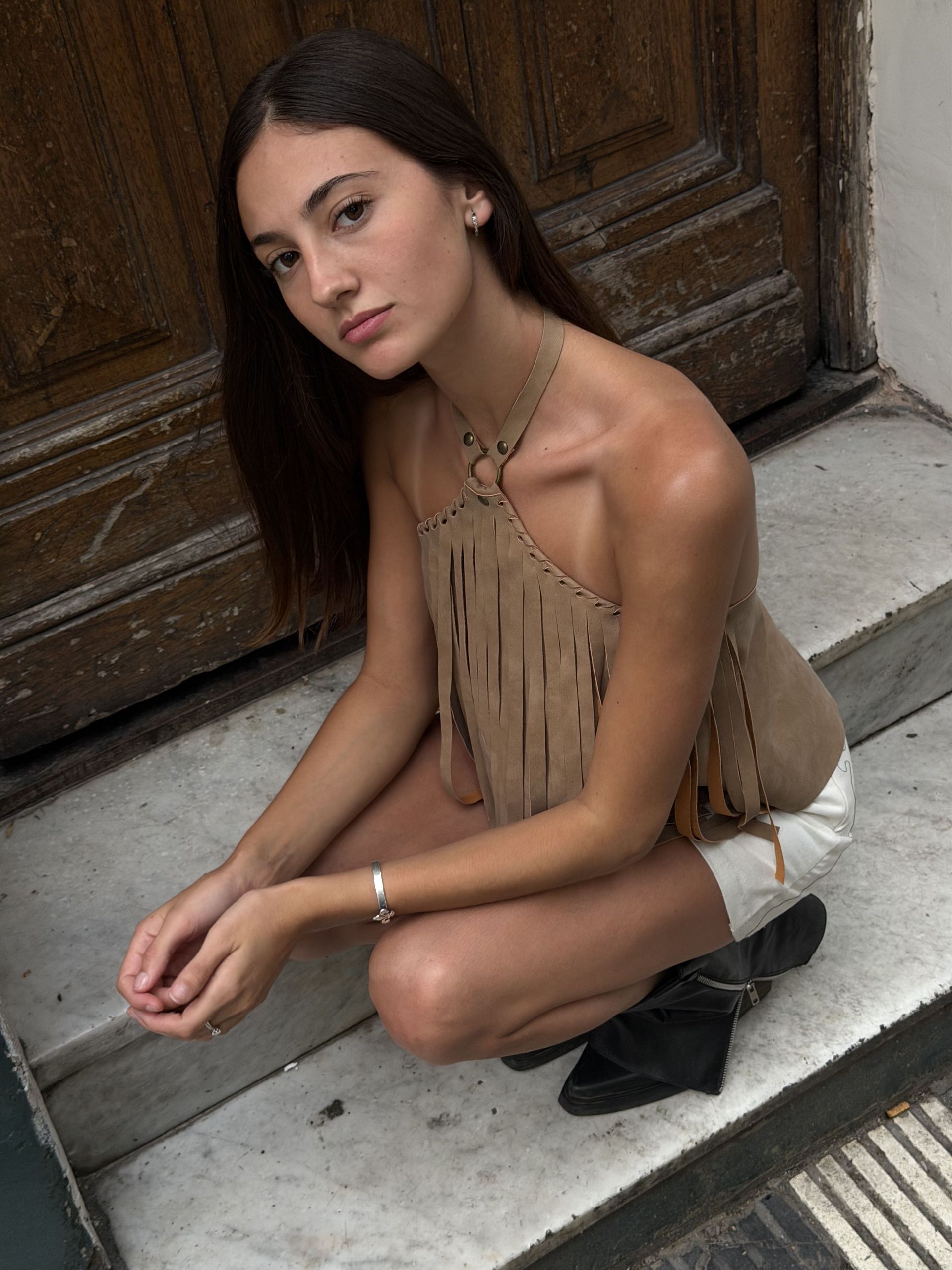 Una mujer se pone en cuclillas en unos escalones vistiendo un top con flecos y pantalones cortos, mirando a la cámara.