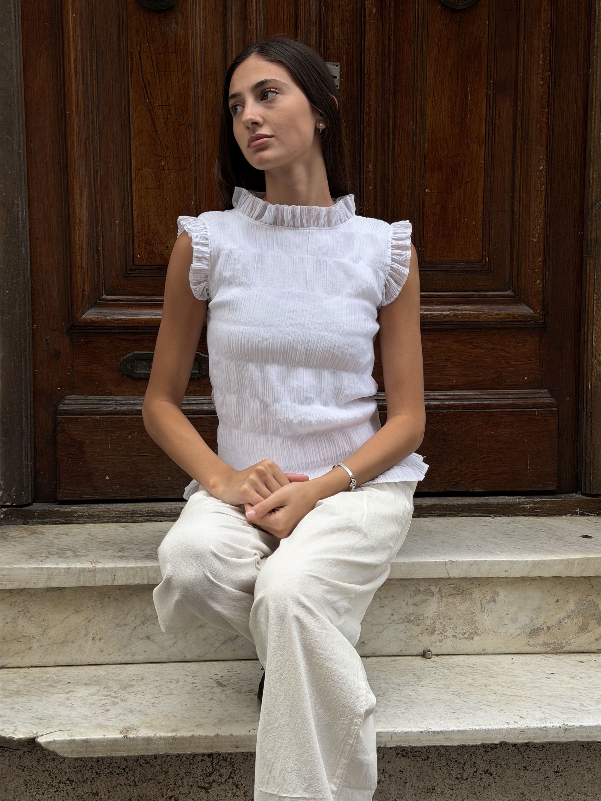 Mujer con top y pantalón blanco, sentada en unos escalones junto a una puerta de madera.