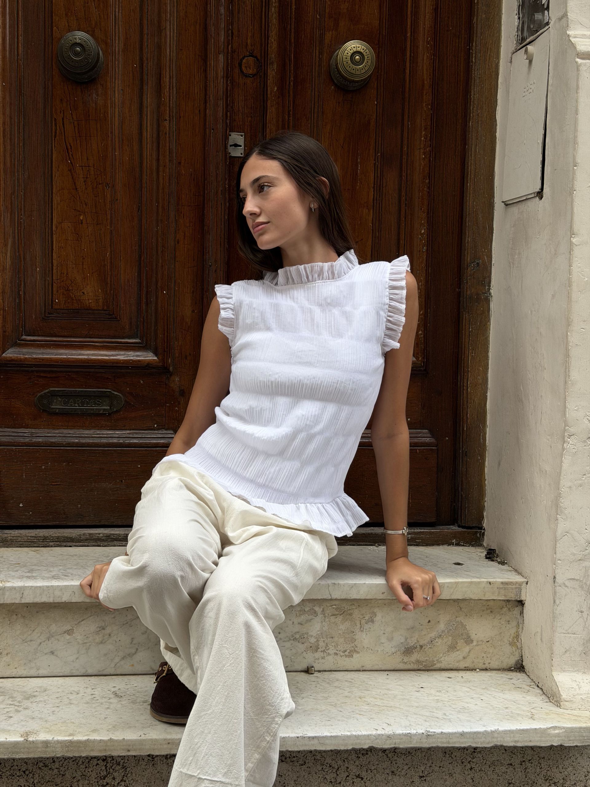 Mujer con top y pantalón blanco, sentada en unos escalones junto a una puerta de madera.