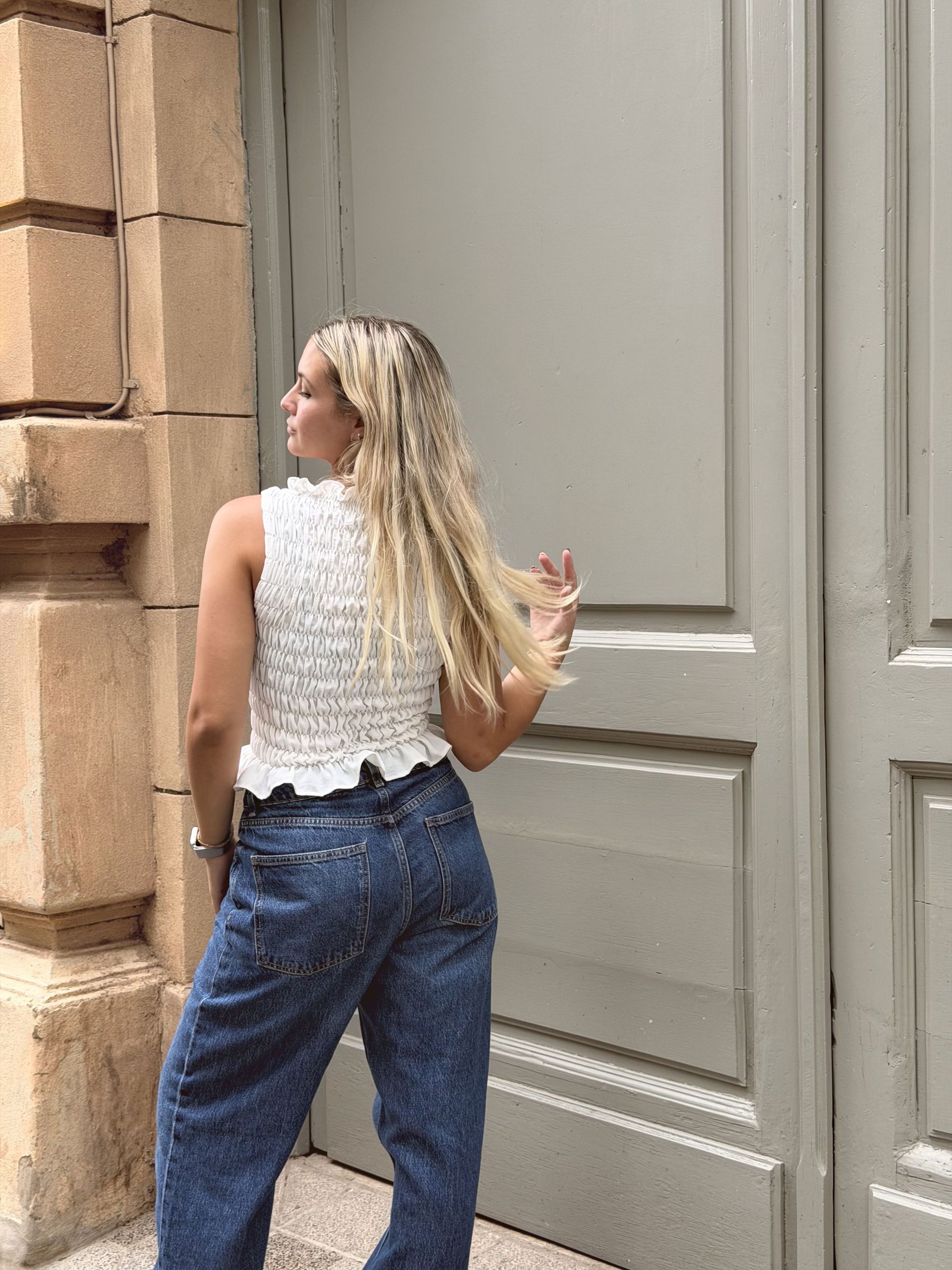 Una mujer con una camiseta blanca y pantalones vaqueros azules posa en una acera frente a una puerta gris.