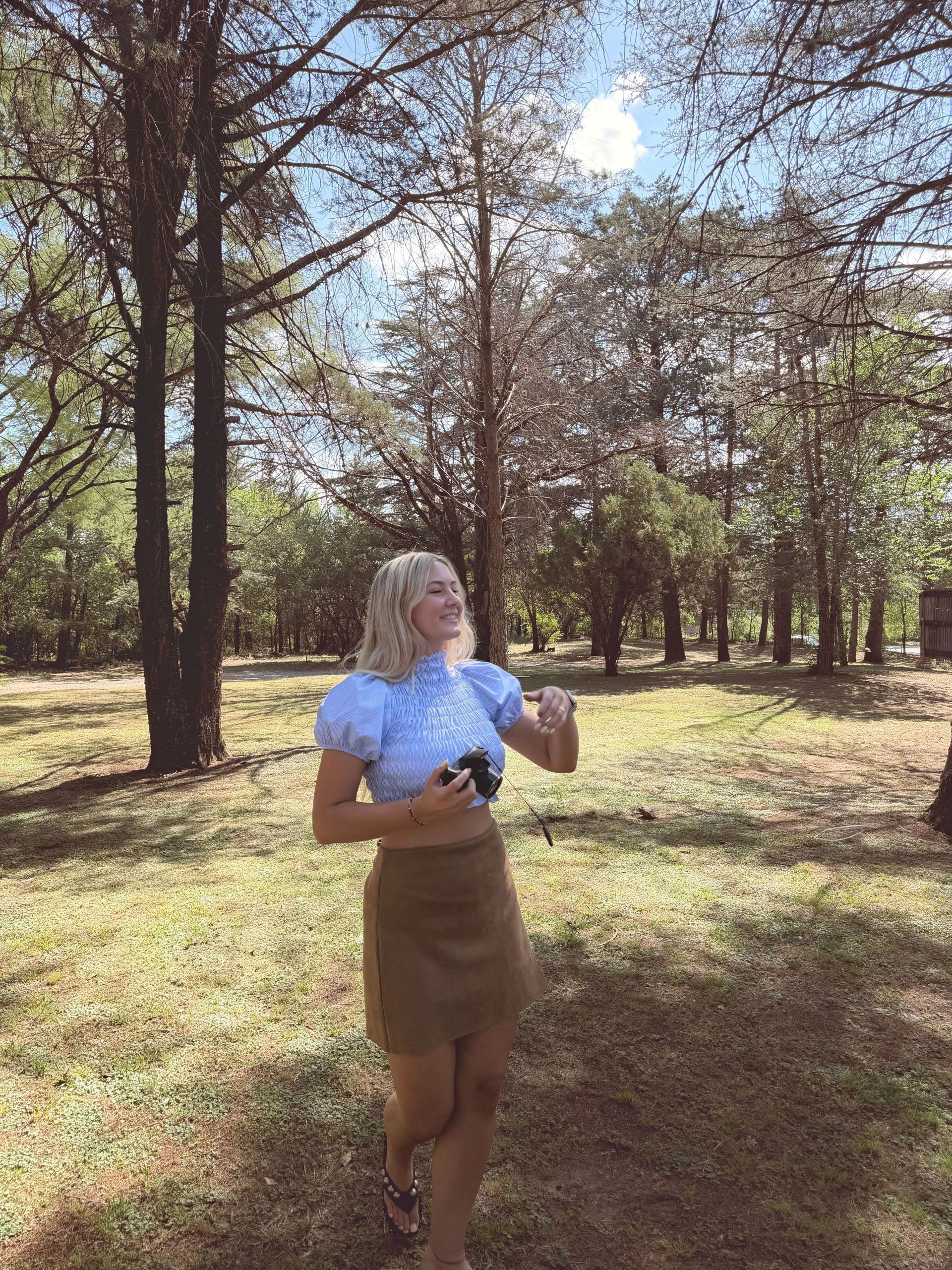 Mujer con top azul y falda marrón sonríe al aire libre, rodeada de árboles.