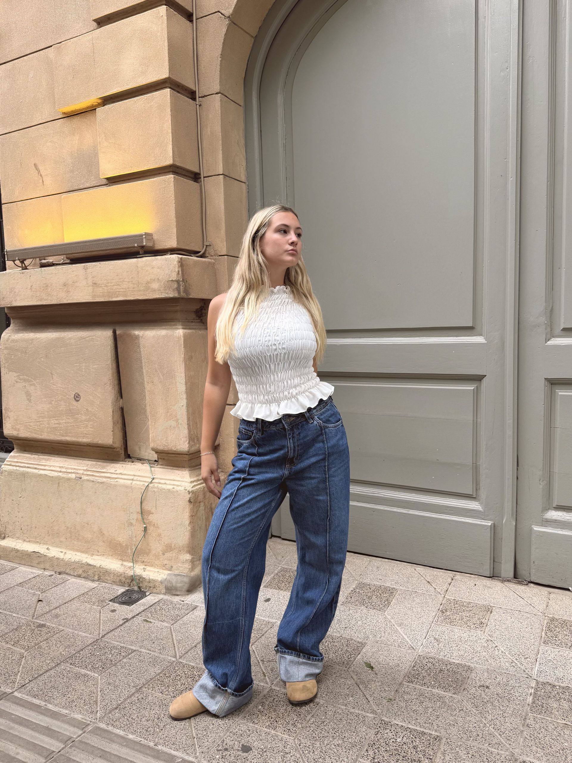 Una mujer con una camiseta blanca y pantalones vaqueros azules posa en una acera frente a una puerta gris.