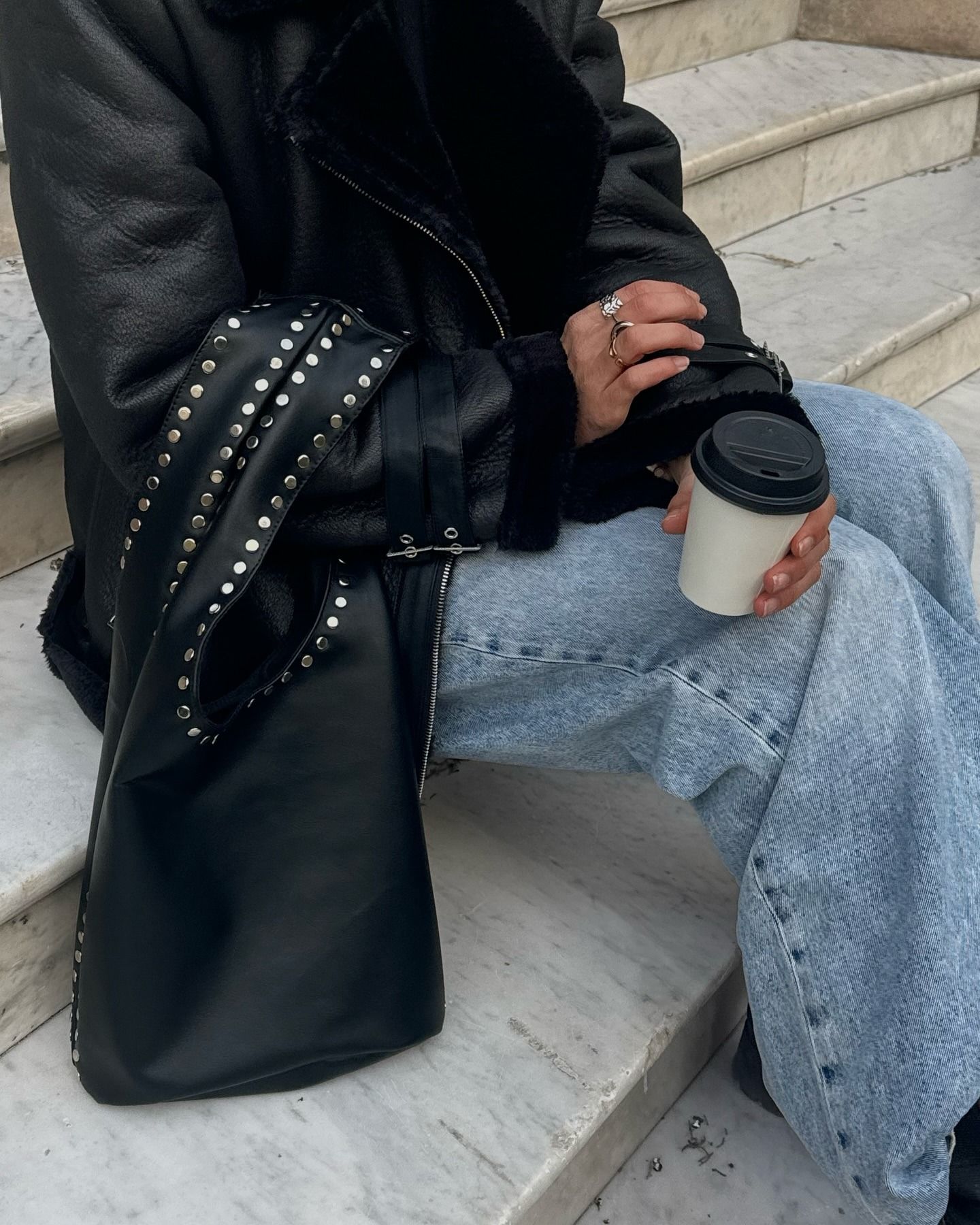 Persona con chaqueta negra y jeans azules, sosteniendo una taza de café, con un bolso negro