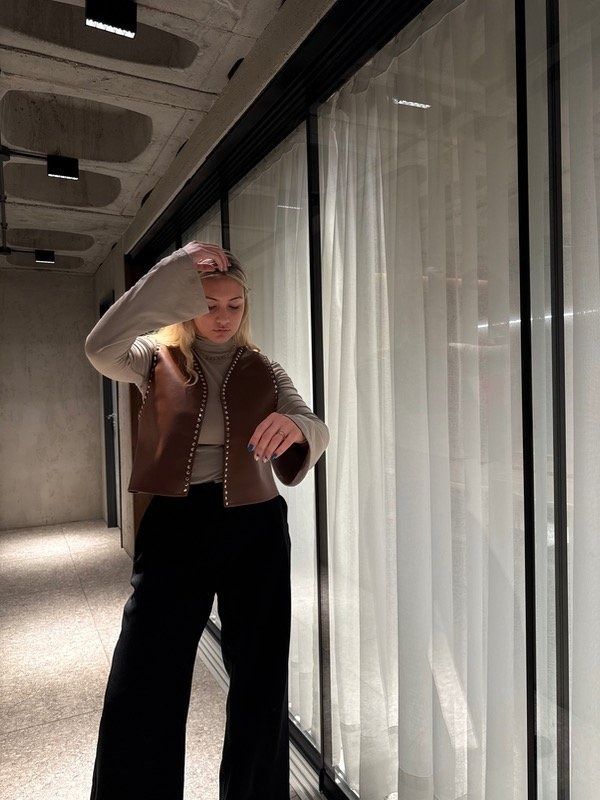 Mujer mirando su reloj en un pasillo, vestida con un chaleco marrón