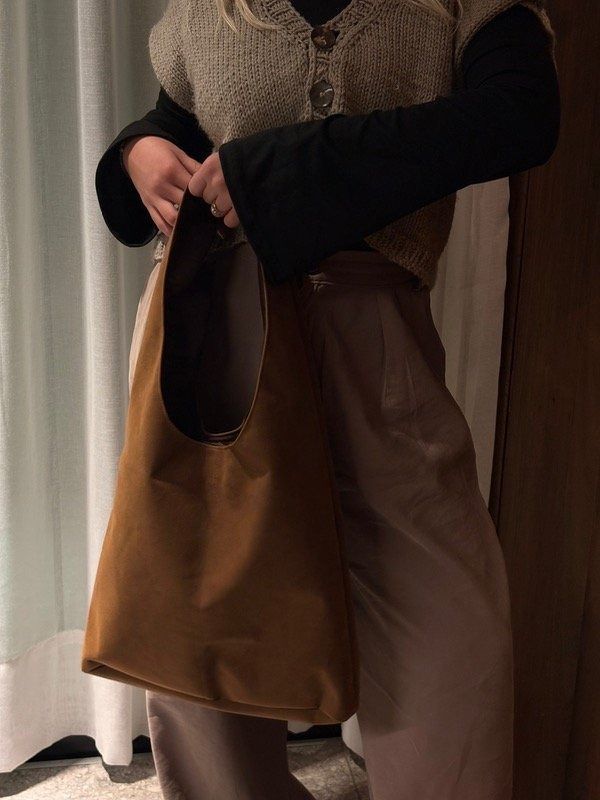 Persona que lleva una bolsa de tela marrón, viste un chaleco de punto y pantalones marrones.