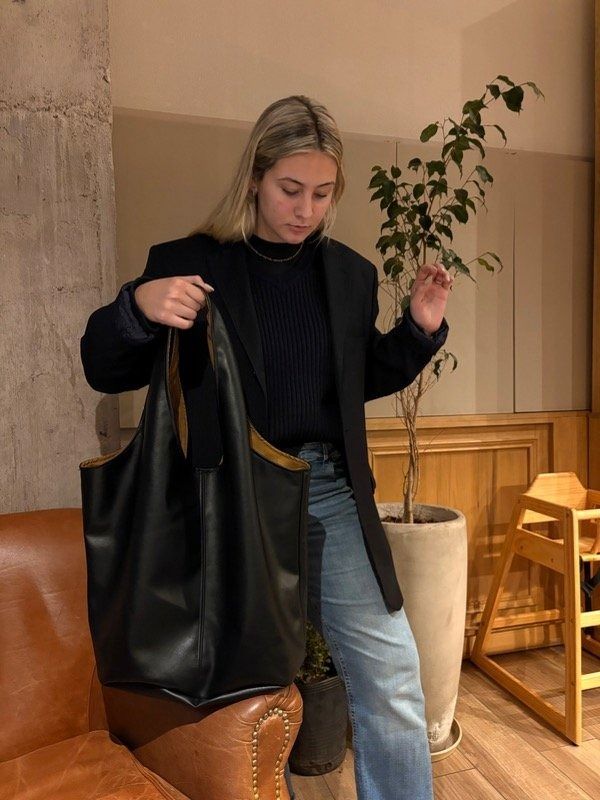 Mujer que sostiene un bolso tote negro grande, vestida con blazer, suéter y jeans en el interior.