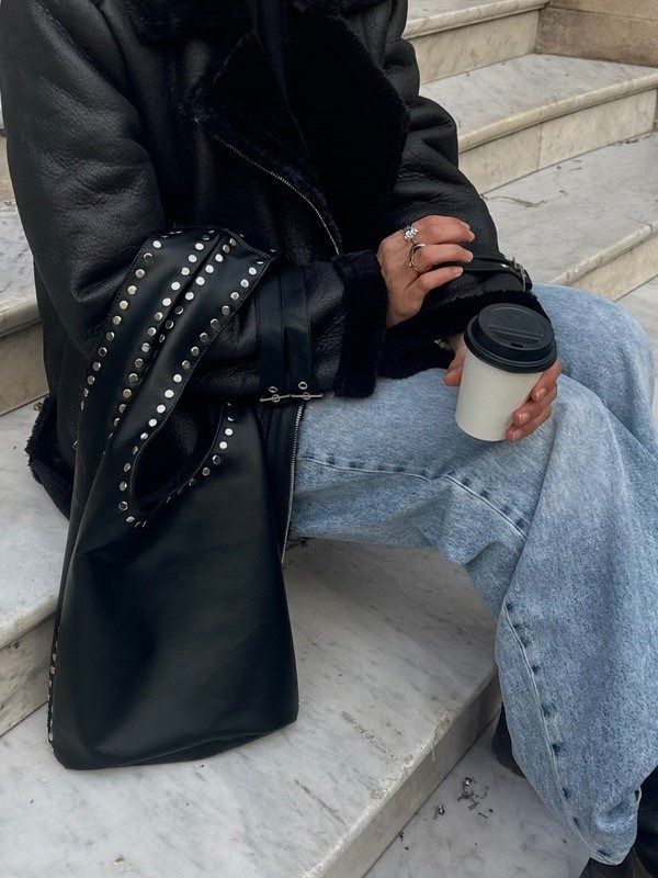 Persona vestida de negro que sostiene un bolso tote con tachuelas, de pie frente a una puerta verde.