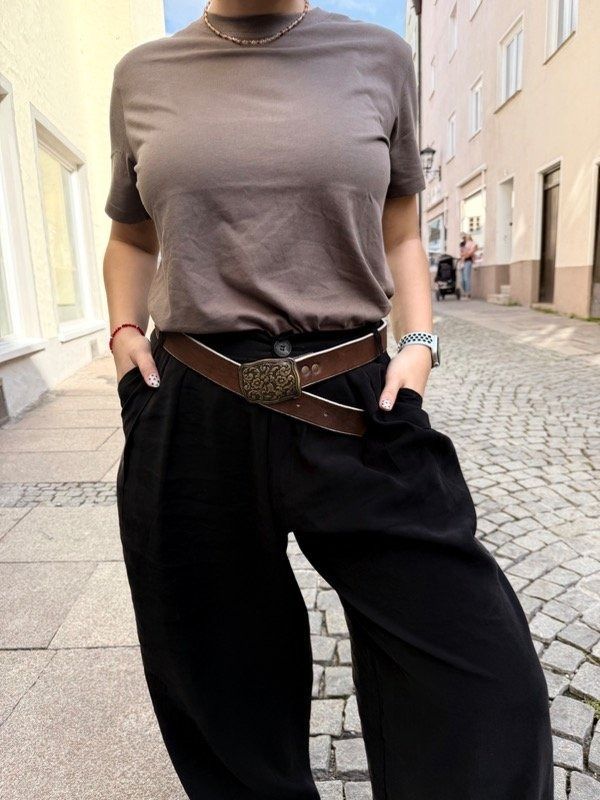 Mujer con blusa marrón y pantalones negros, manos en los bolsillos, con un cinturón marrón en una calle empedrada.