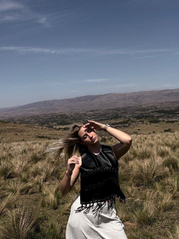 Mujer protegiéndose los ojos, de pie en un campo con una cordillera al fondo, cielo azul.
