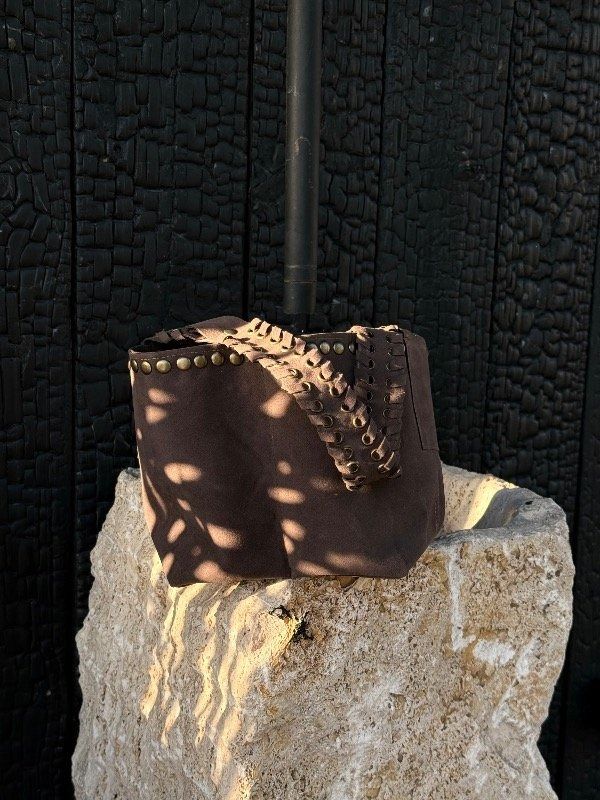 Bolso de cuero marrón sobre un bloque de piedra tosca, contra una pared de madera negra.