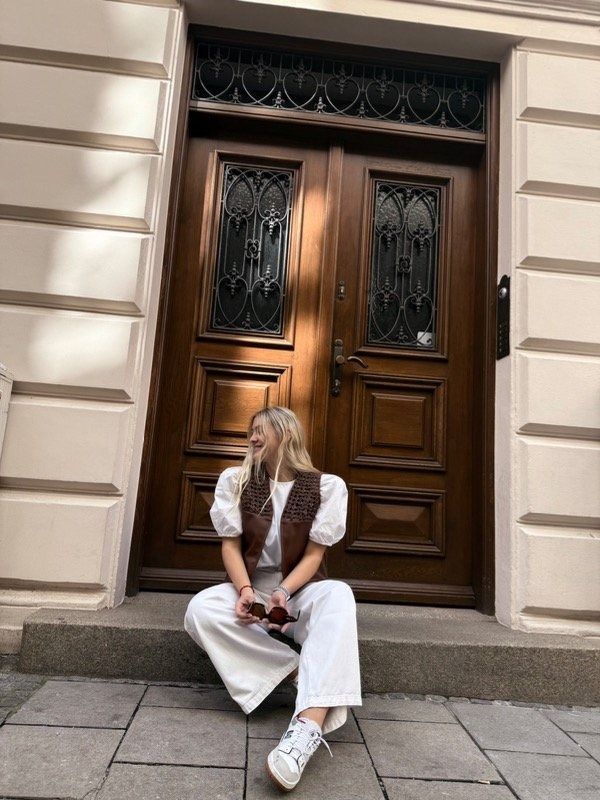 Una mujer con chaleco marrón y blusa blanca sonríe, con la mano en la cabeza, delante de una puerta de madera.