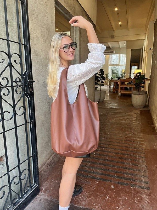 Una mujer con blusa blanca y bolso marrón levanta el brazo, sonriendo, en un pasillo de ladrillo.