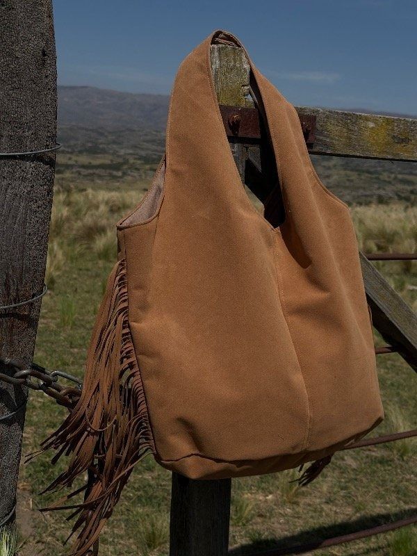 Bolso marrón de ante con flecos colgado de una valla de madera, al aire libre.